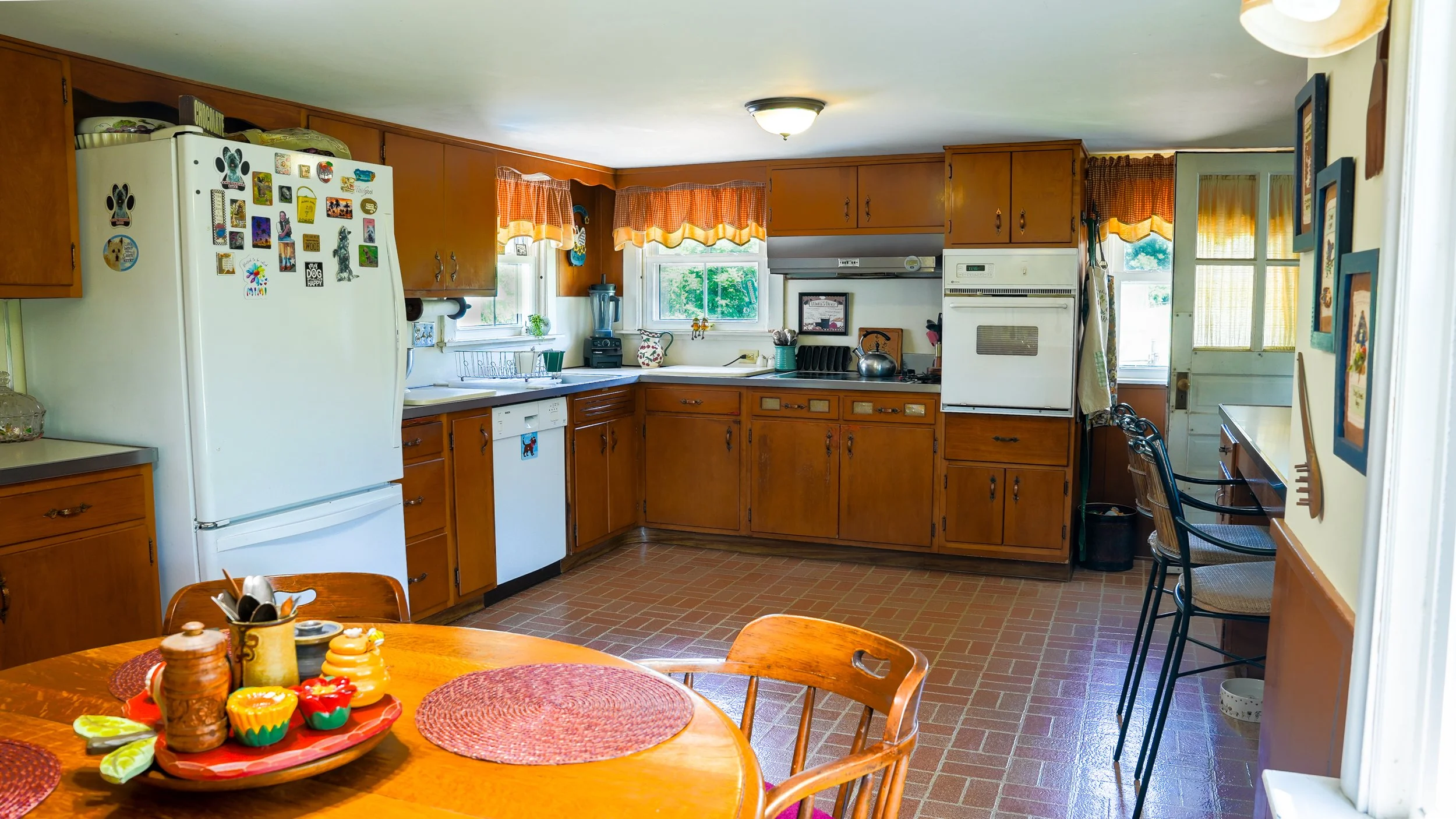 A kitchen with wooden cabinets, a white refrigerator decorated with magnets, and a window with orange checkered curtains. There is a blender on the counter, a stovetop, and a door leading outside. A table with a plate of cupcakes and utensils is in the foreground.
