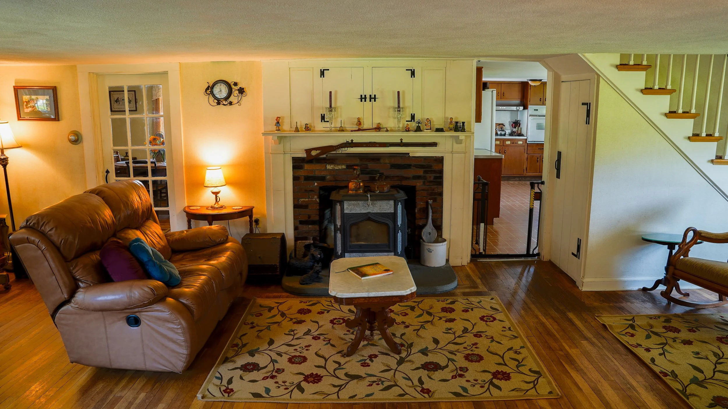 Living room with leather couch, fireplace, and wooden floors, decorated with lamps, framed pictures, and a colorful rug.
