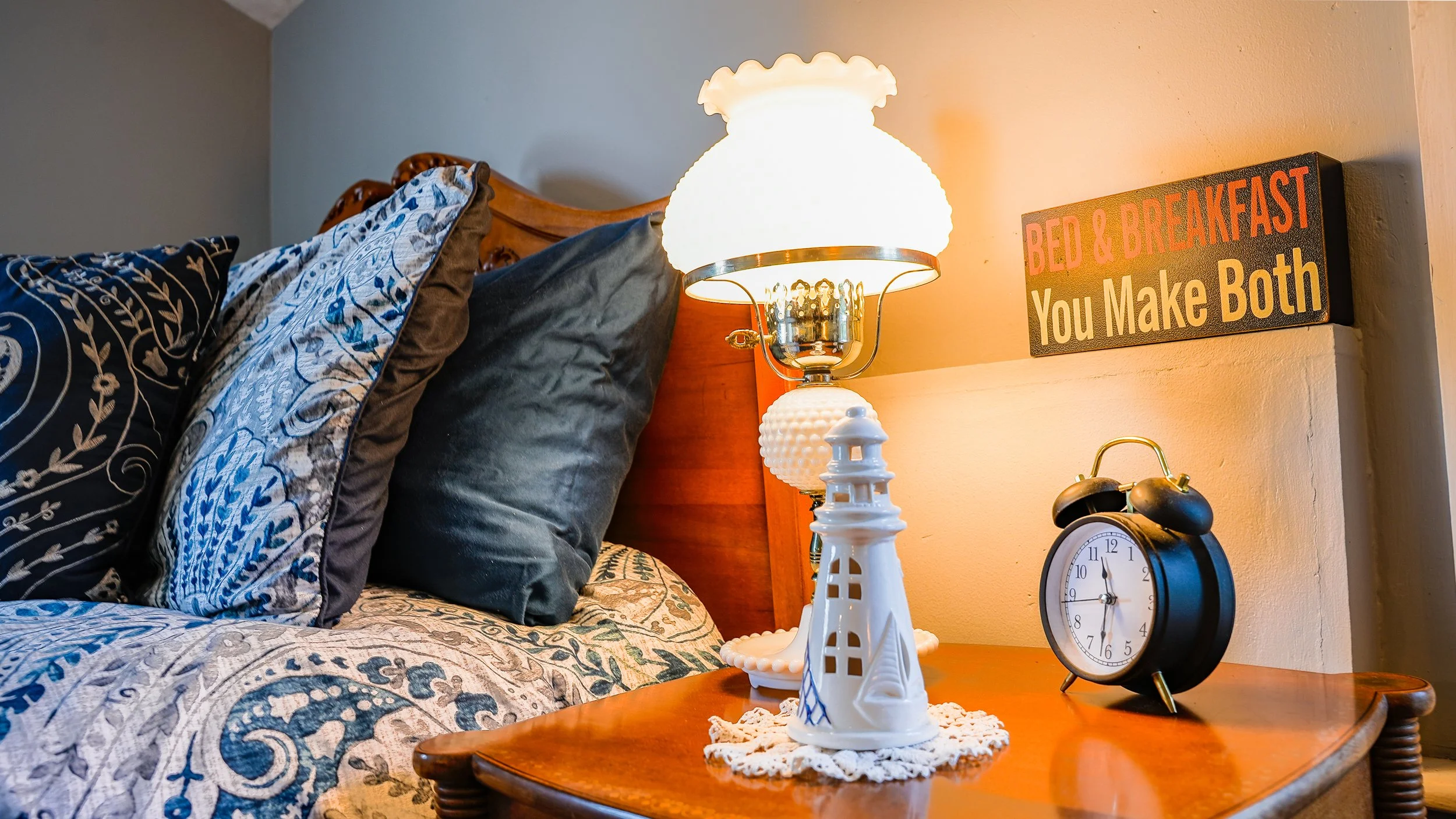 Close-up of a bedroom nightstand with a lighthouse-shaped lamp, a black alarm clock, and a small white lighthouse decor piece. Behind the lamp, a bed with patterned and solid pillows is visible, and a sign on the wall reads, 'BED & BREAKFAST You Make Both'.