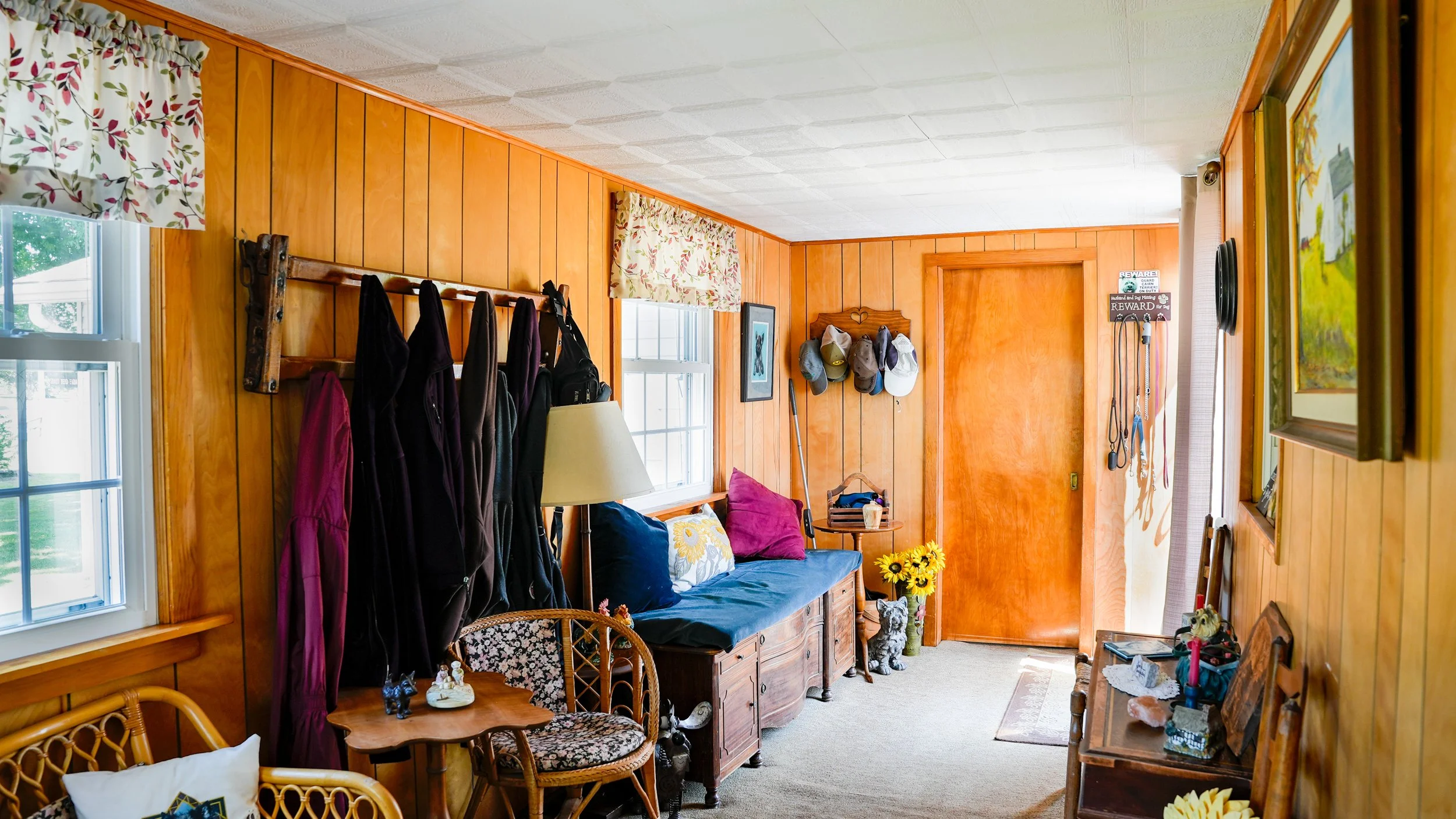 A cozy living room with wood-paneled walls in a mid-century style, featuring windows with floral curtains, a blue couch with colorful pillows, a wooden side table with sunflowers, and various decorations including hats hanging on the wall and framed pictures.
