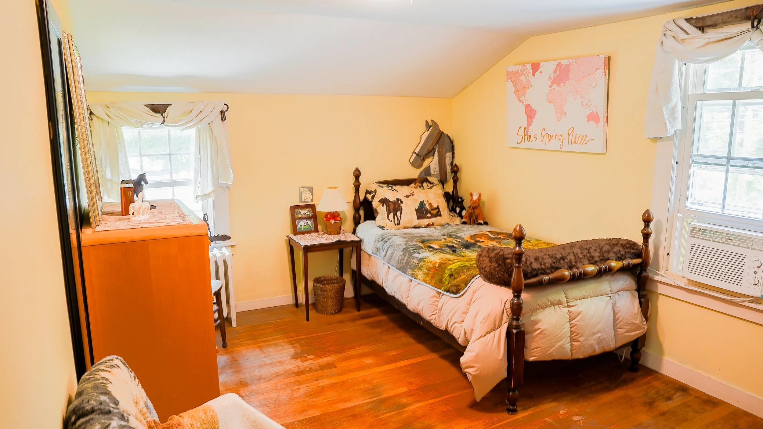 A cozy bedroom with a single bed featuring animal-themed bedding, a small wooden table with a lamp, a framed picture, and a stuffed animal. There's a window with white curtains, a wall art piece with a world map and the words 'She's Going Places,' and a window air conditioning unit. The room has wooden floors and soft yellow walls.