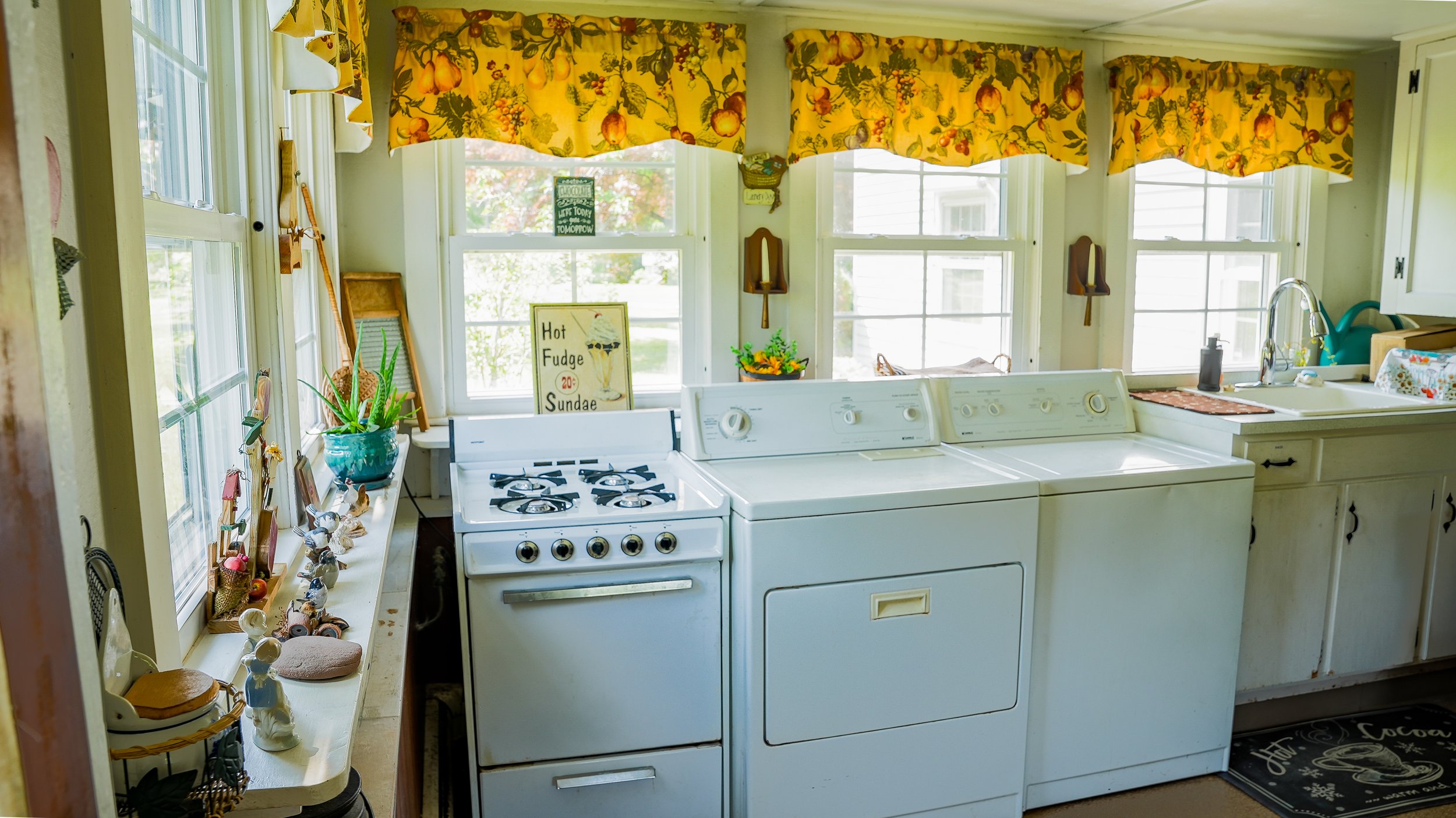 Kitchen with yellow floral curtains, white appliances, and decorated windowsill with plants and trinkets.