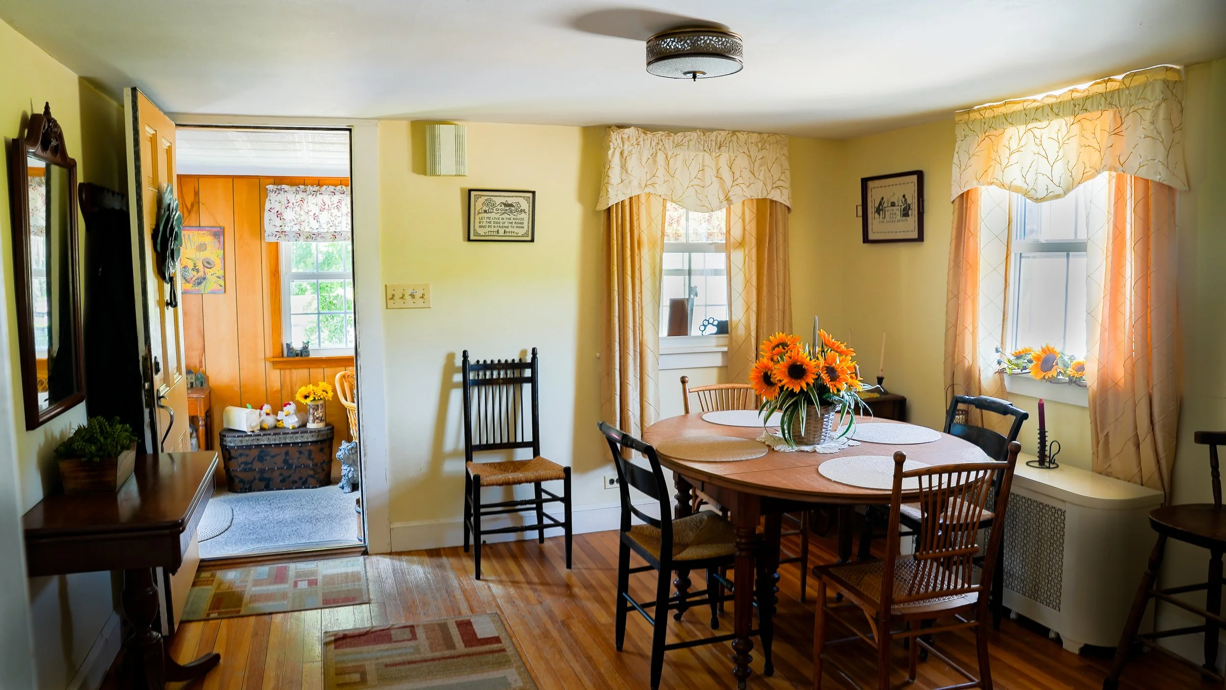 A cozy dining room with a wooden dining table and chairs, decorated with a sunflower centerpiece, located in a yellow-painted room with windows covered by cream and peach curtains, and framed pictures on the walls.