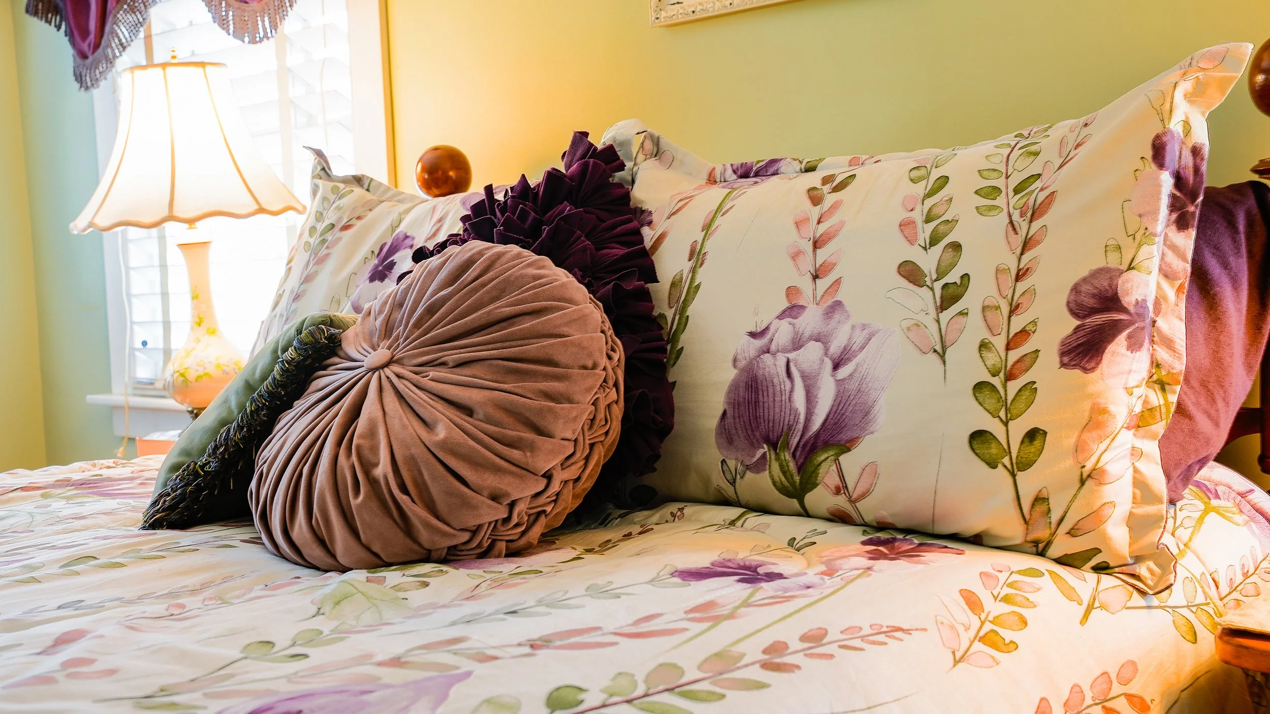 A bed with floral patterned bedding and decorative pillows, including a large round pillow and a fan-shaped pillow, in a cozy, yellow-walled bedroom with a lamp and window in the background.