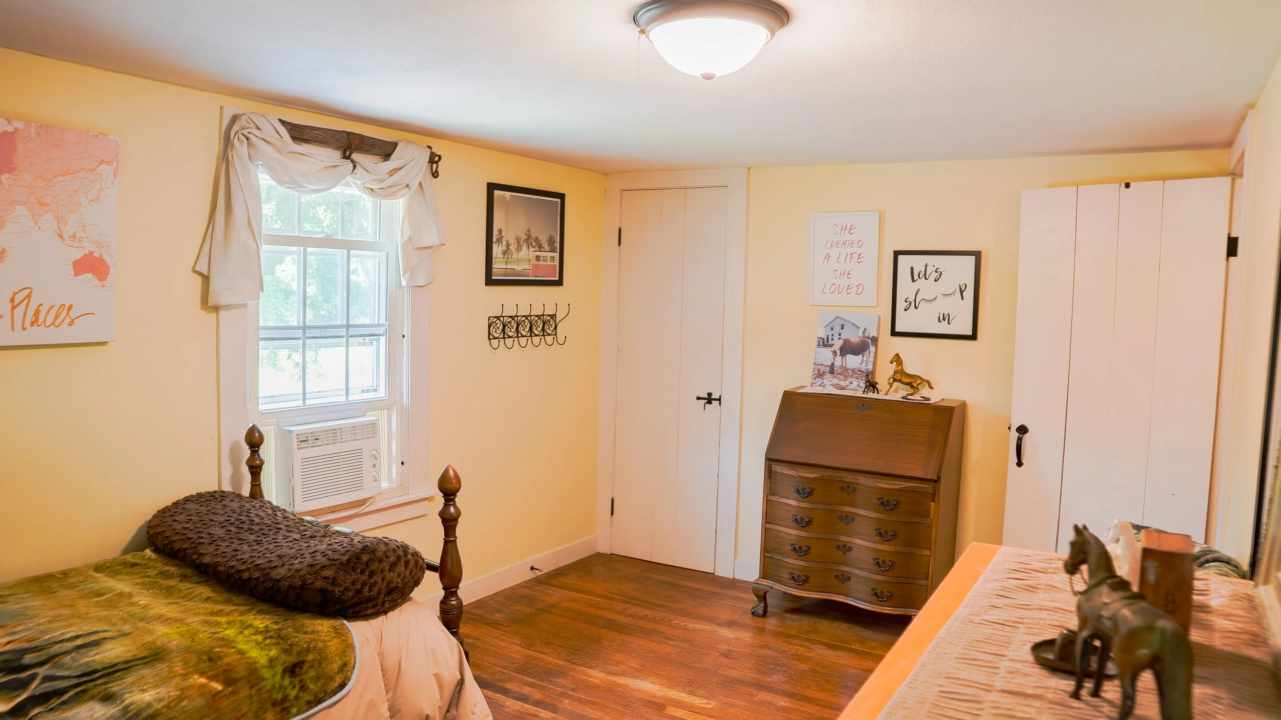A cozy bedroom with a yellow wall, a window with white curtains, and a bed with a green and brown blanket. There are framed pictures and decorative items on the wall and a wooden dresser with small horse figurines on top.