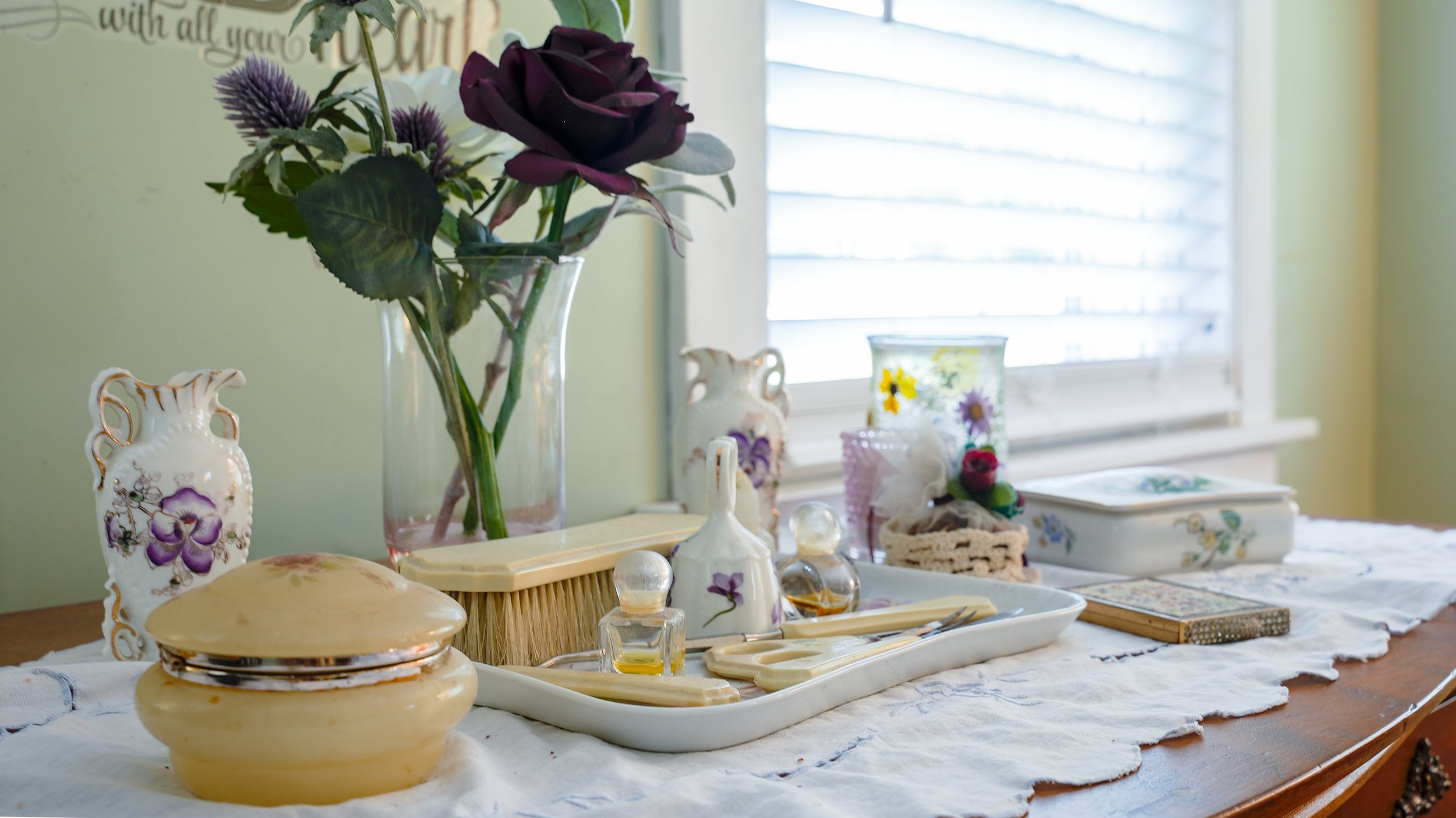 A table decorated with vintage and floral items, including a bouquet of dark purple roses in a glass vase, ceramic vases with purple and yellow flowers, a basket, and various small decorative objects, set near a window with blinds.