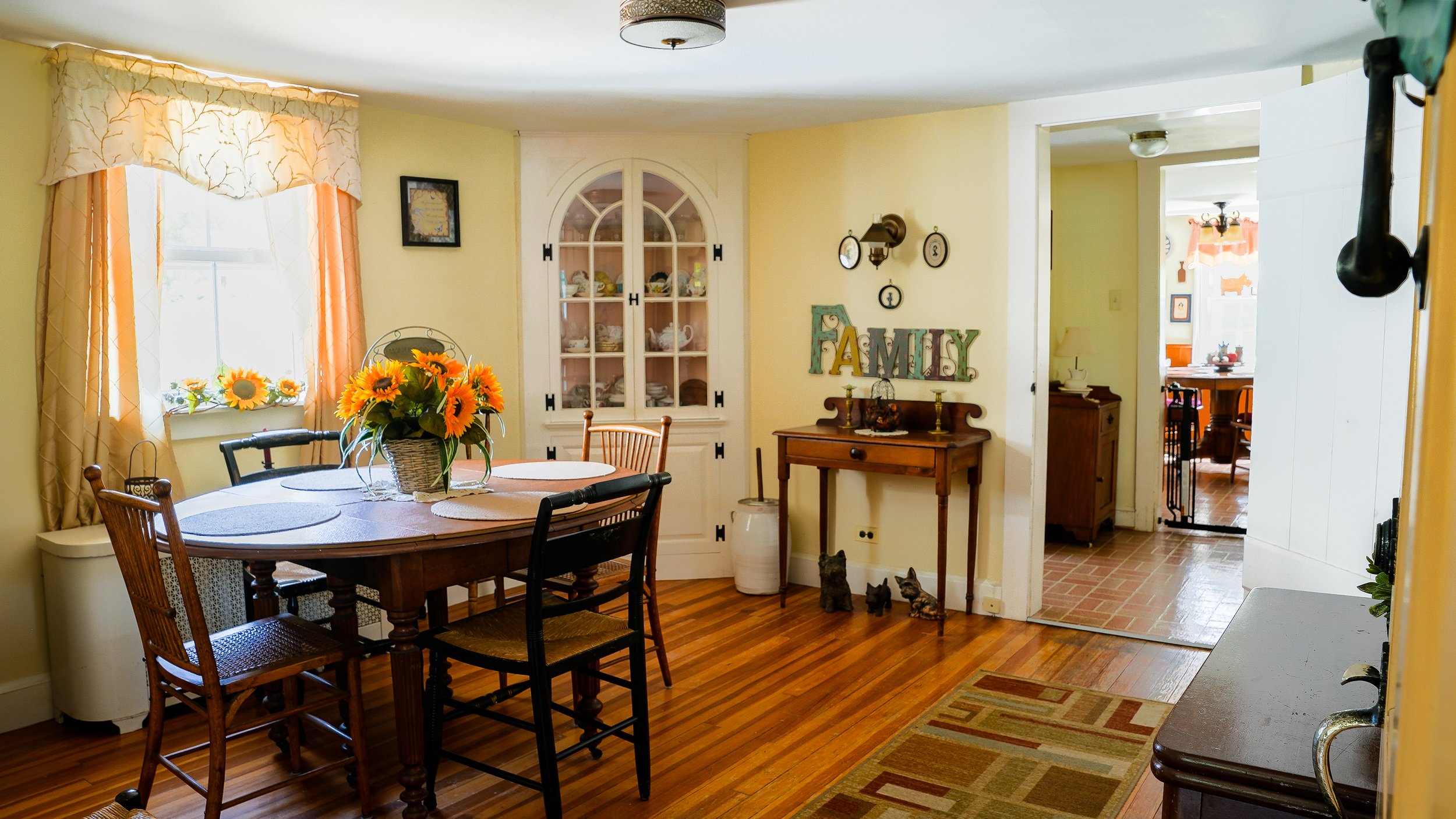 A cozy dining area with a wooden table, sunflower centerpiece, and mixed chairs near a window with beige curtains. The wall features decorative plates and a sign that reads 'FAMILY'. There's a glass-fronted cabinet displaying dishware, and a doorway leading to another room with a brick floor and a wooden dining set. Small cat figurines are on the floor near a small table.