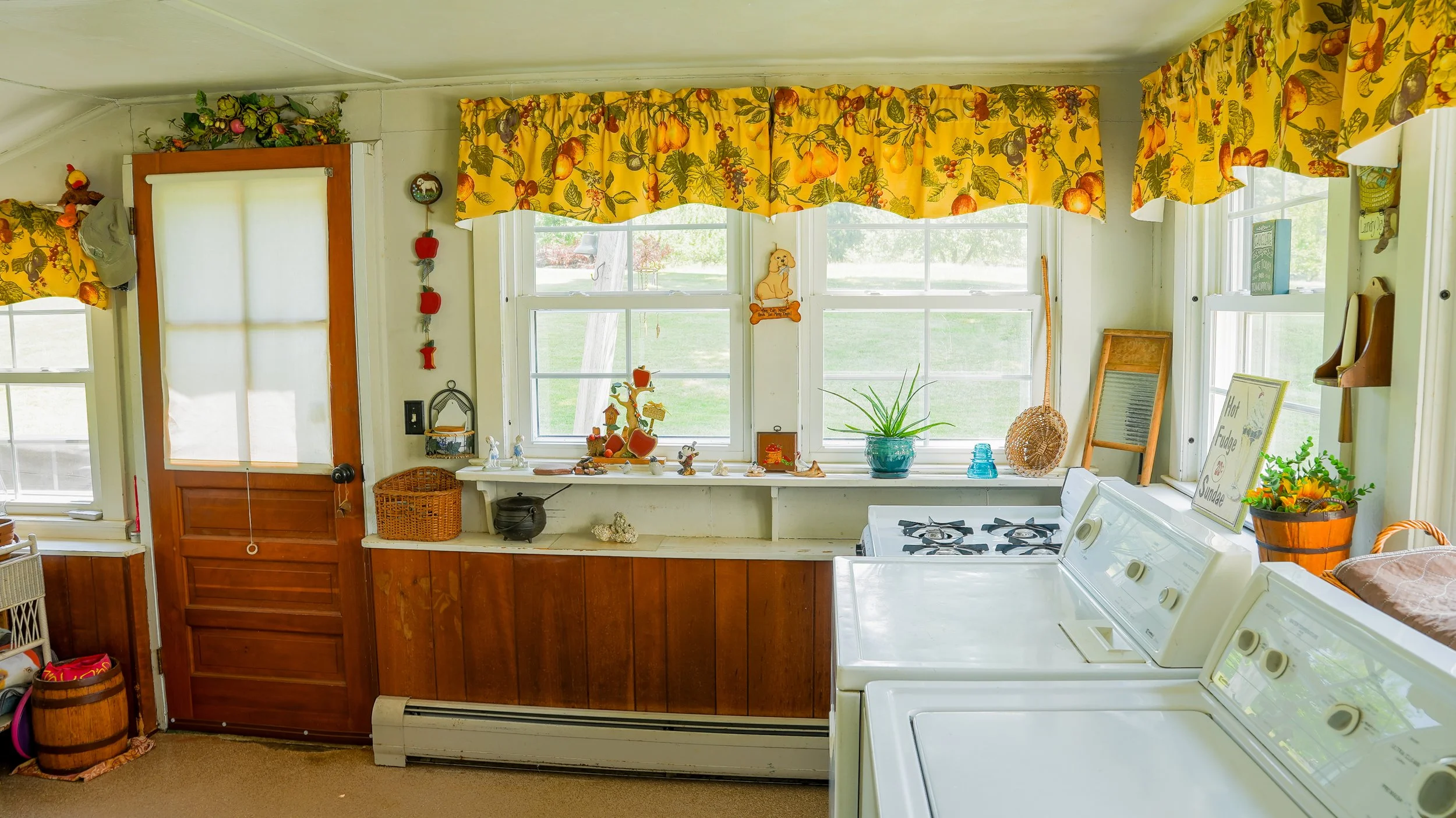 A cozy kitchen with a row of large windows topped with yellow fruit-patterned valances, decorated with various figurines, plants, and knick-knacks on the windowsills and walls, and white appliances including a washing machine and dryer.