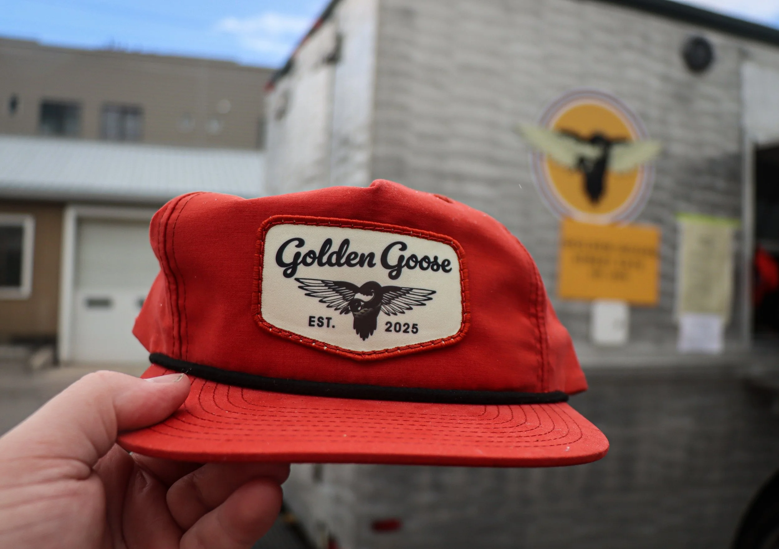 A red cap with a 'Golden Goose' logo being held up by a person's hand in front of a building with a large  bird logo and a sign.