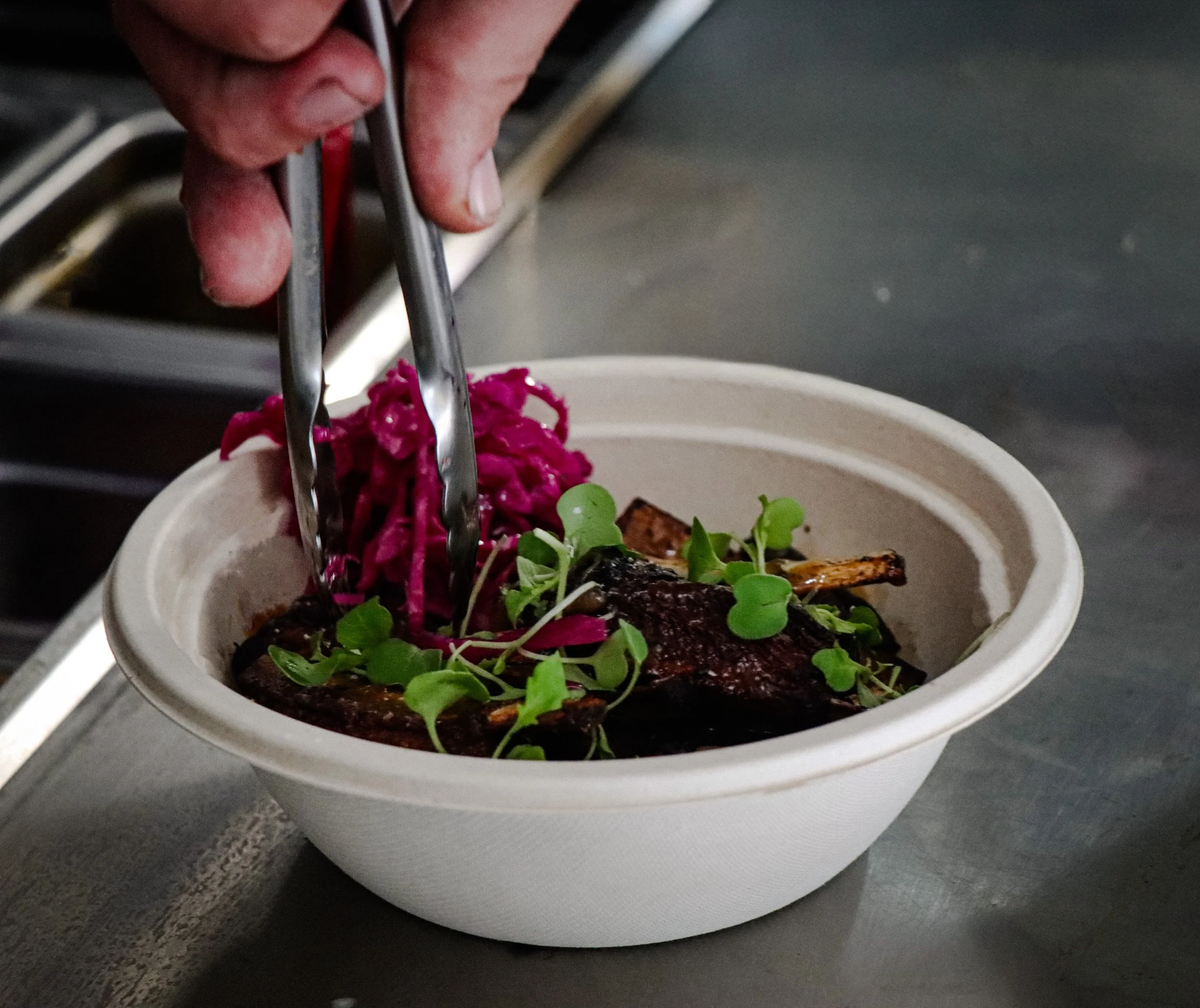 Close-up of a hand holding tongs and placing food into a slow cooker. The food appears to be meat with some vegetables or garnish on top. The background shows a dark kitchen counter.