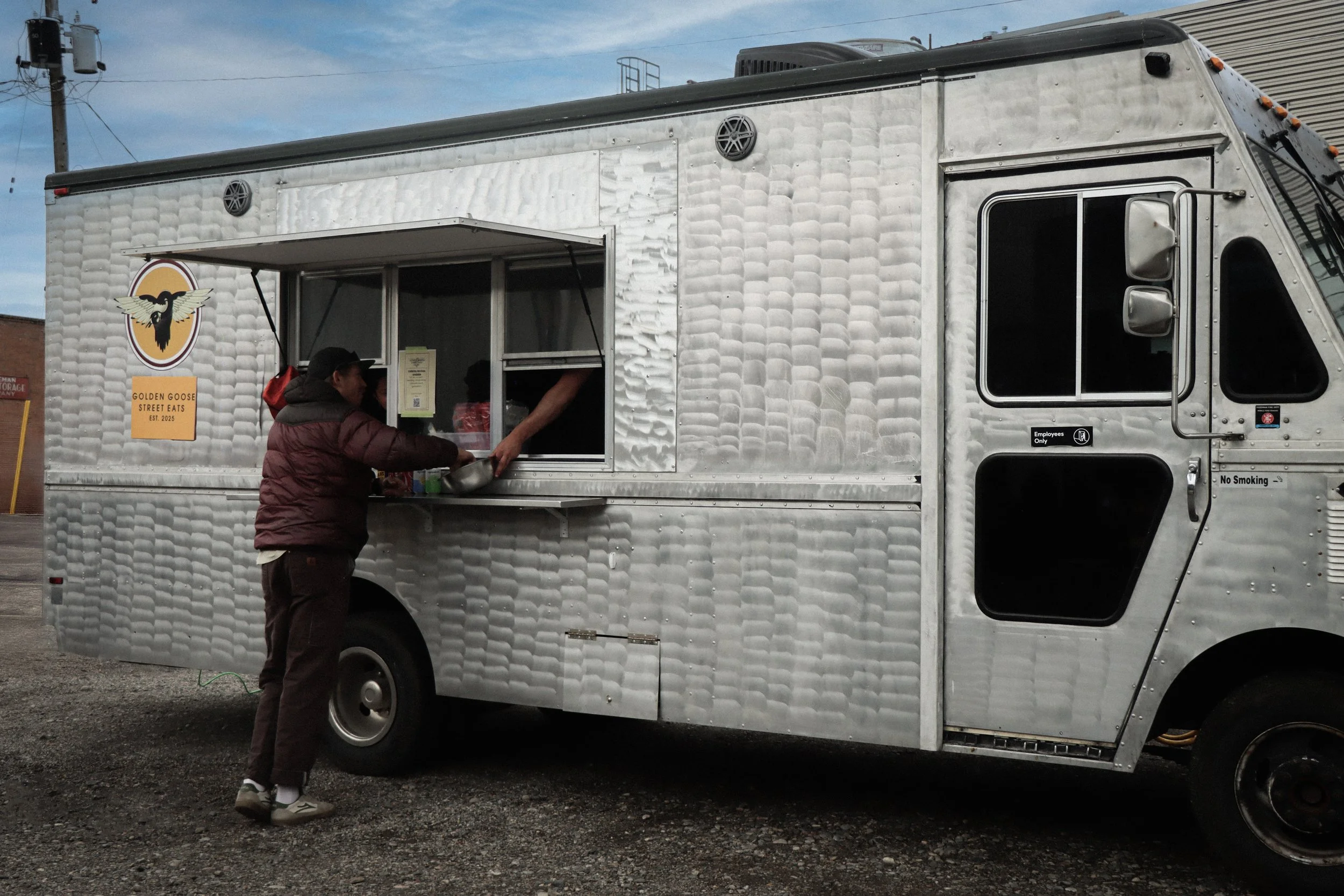 A food truck with a silver, textured exterior, a serving window with a person inside, and a customer placing an order. The truck has a logo featuring a black bird with wings spread, and a sign that reads 'Golden Goose Street Eats Est. 2025.'