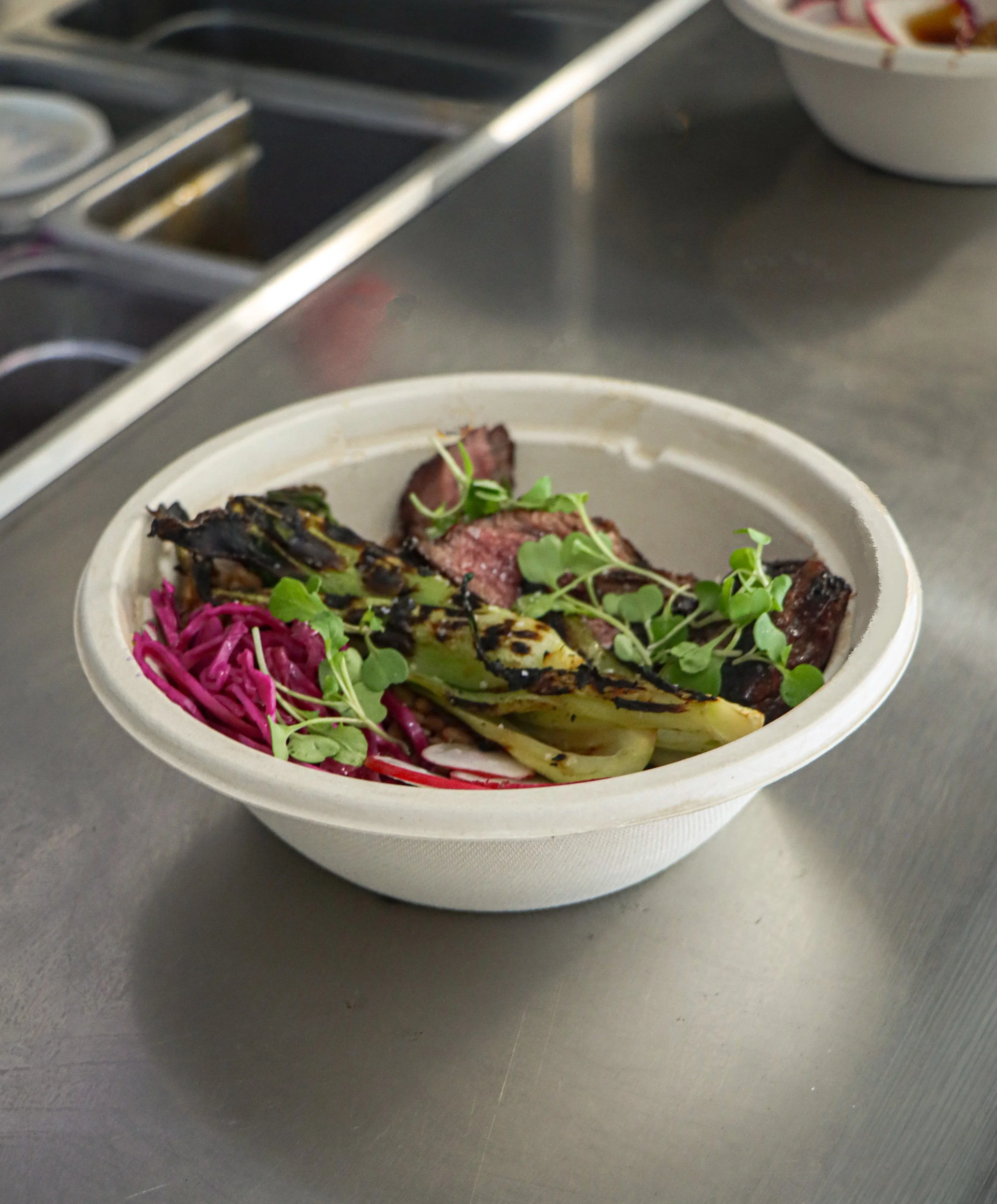 A bowl of food with grilled meat, purple shredded vegetables, and green microgreens on a metallic surface.
