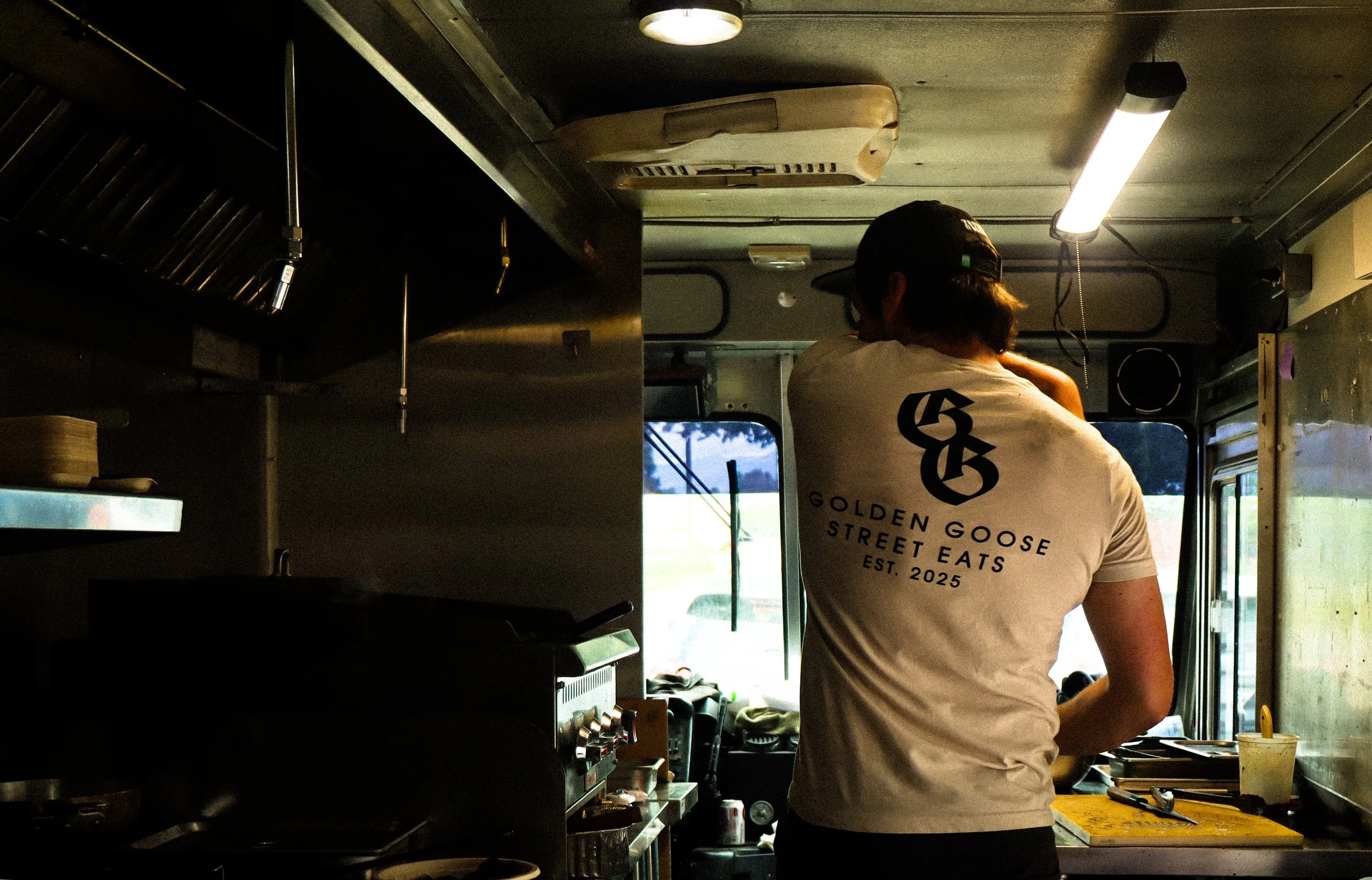 A person working inside a food truck, wearing a white t-shirt with the logo 'Golden Goose Street Eats' and standing near a counter with a yellow cutting board and utensils, with kitchen equipment visible around.