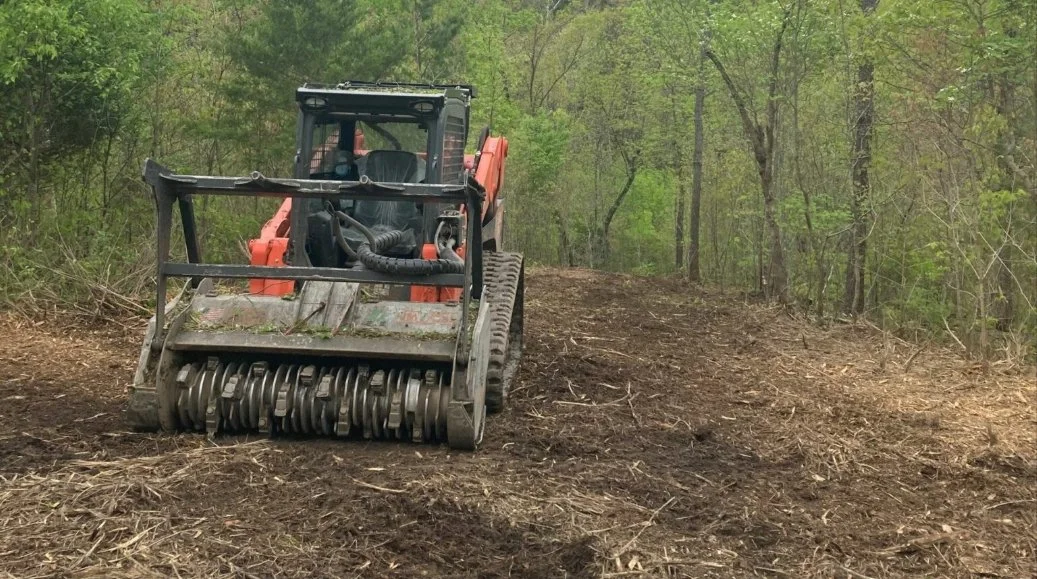 A small orange and black construction vehicle with a large roll of metal blades, working on clearing a dirt path through a wooded area with green trees.
