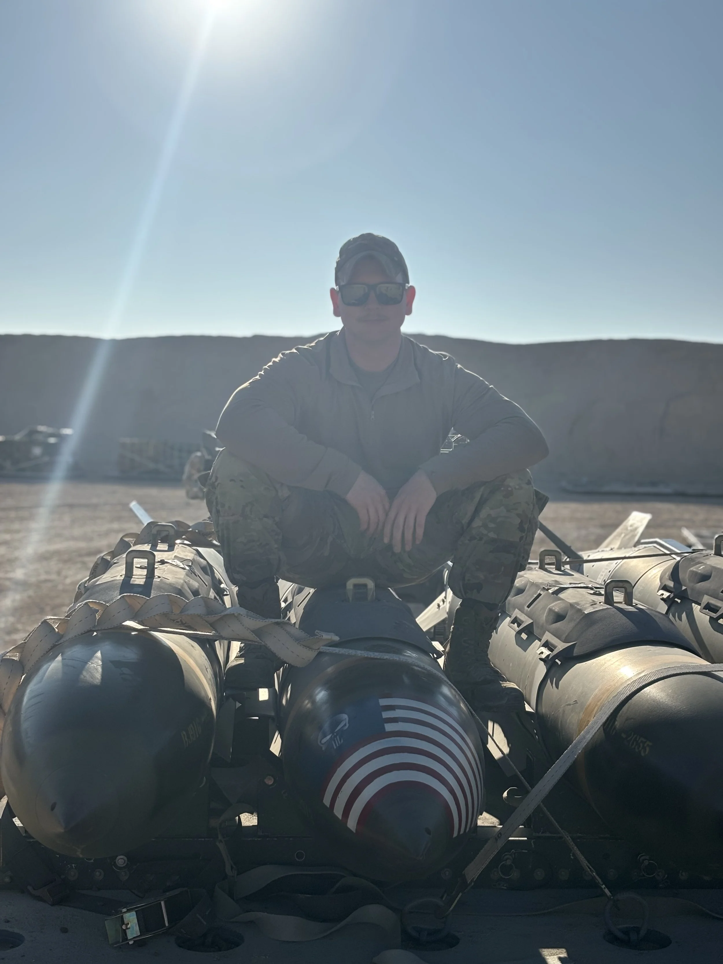 The owner sitting on top of stacked military bombs, wearing camouflage pants, a hoodie, sunglasses, and a cap, in a desert landscape with mountains in the distance and the sun shining brightly overhead.