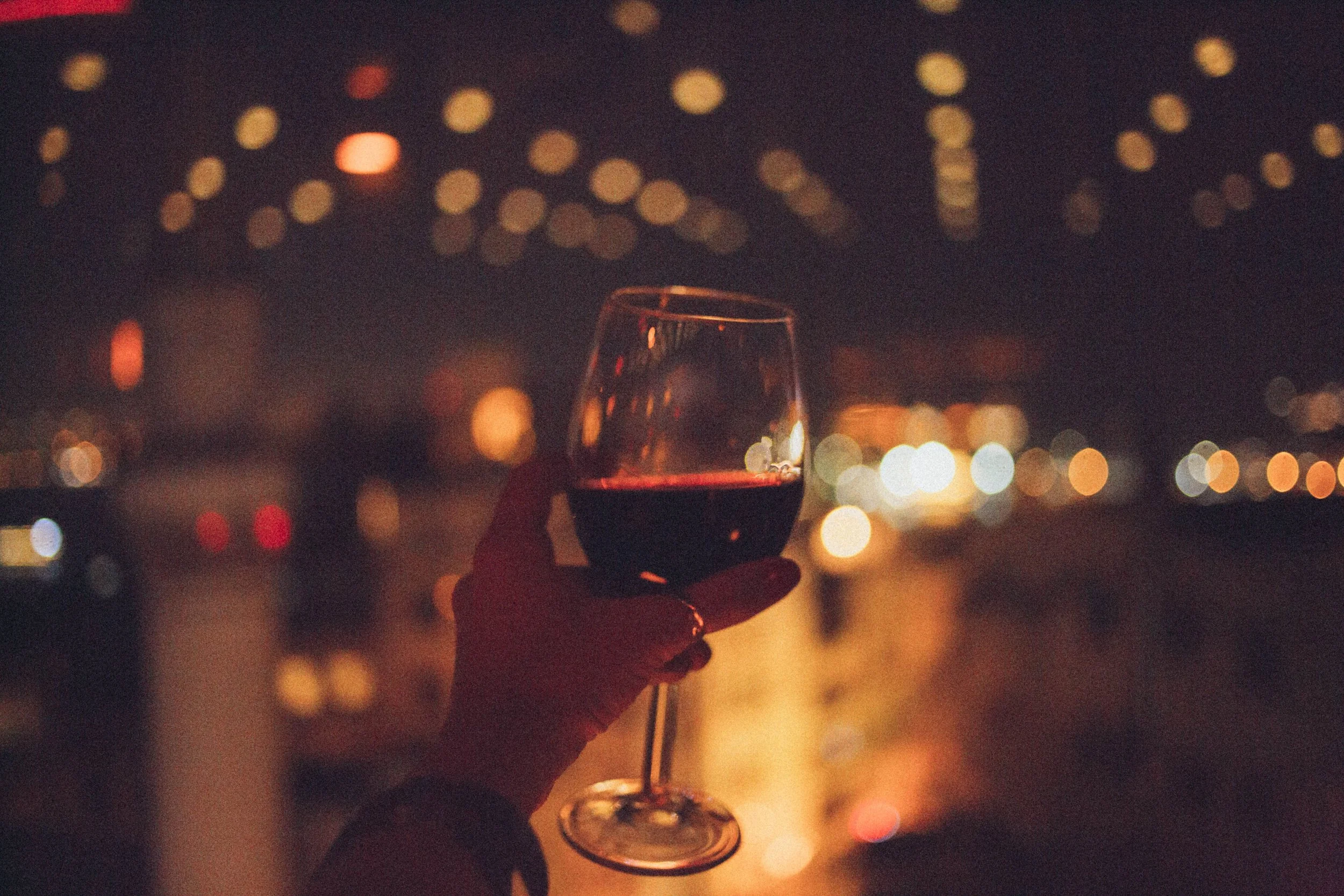 A glass of red wine held against a softly lit downtown backdrop, reflecting the warm, inviting atmosphere of Palace Wine Bar in Rockford, Illinois.