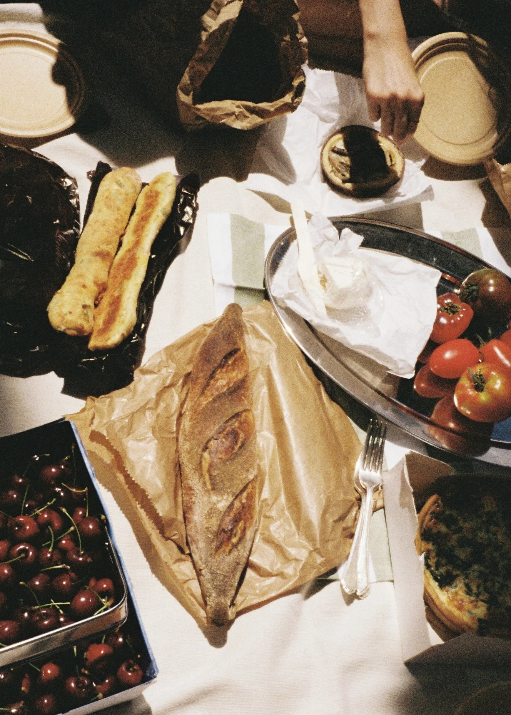 A table with various baked goods including a long loaf of bread, two breadsticks, a tart with a dark filling, and a slice of pizza. There are also containers of cherries, tomatoes, and other assorted food items.