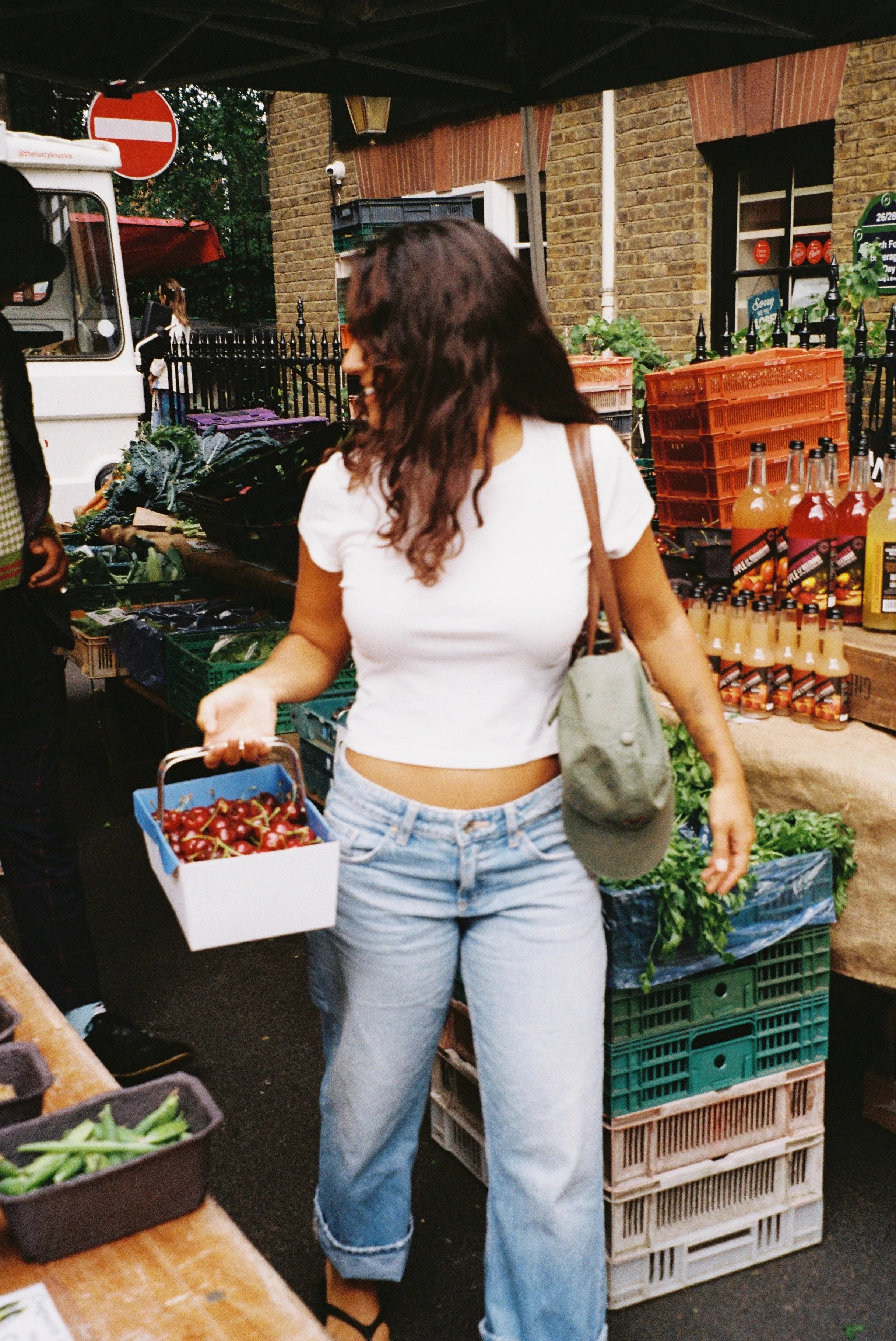 Woman shopping at an outdoor farmer's market, holding a box of cherries, surrounded by various fresh vegetables and fruits on display.