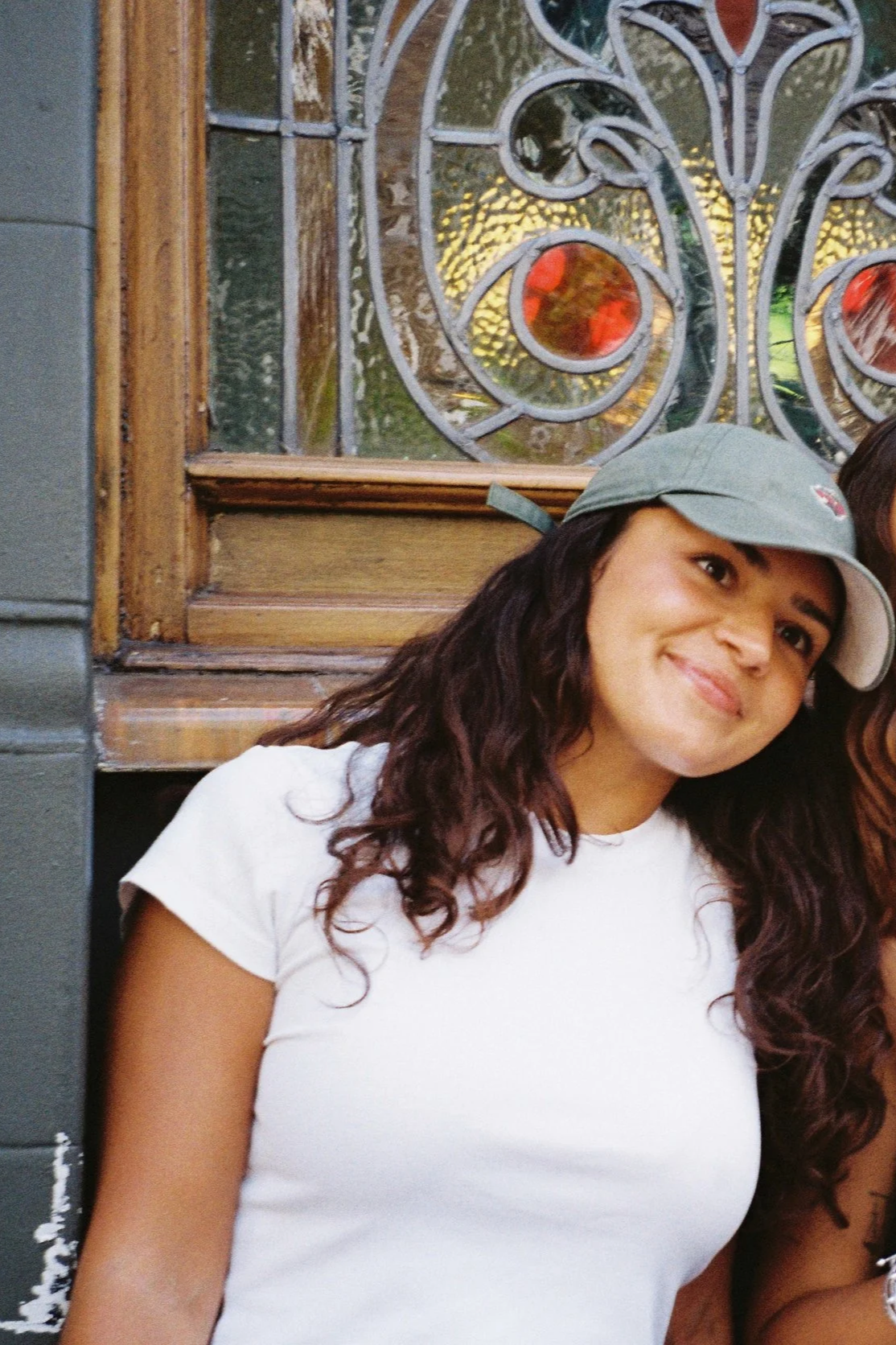 A woman with long dark hair smiling and wearing a white tank top and light gray pants stands outdoors in front of a stained glass window with a green cap.