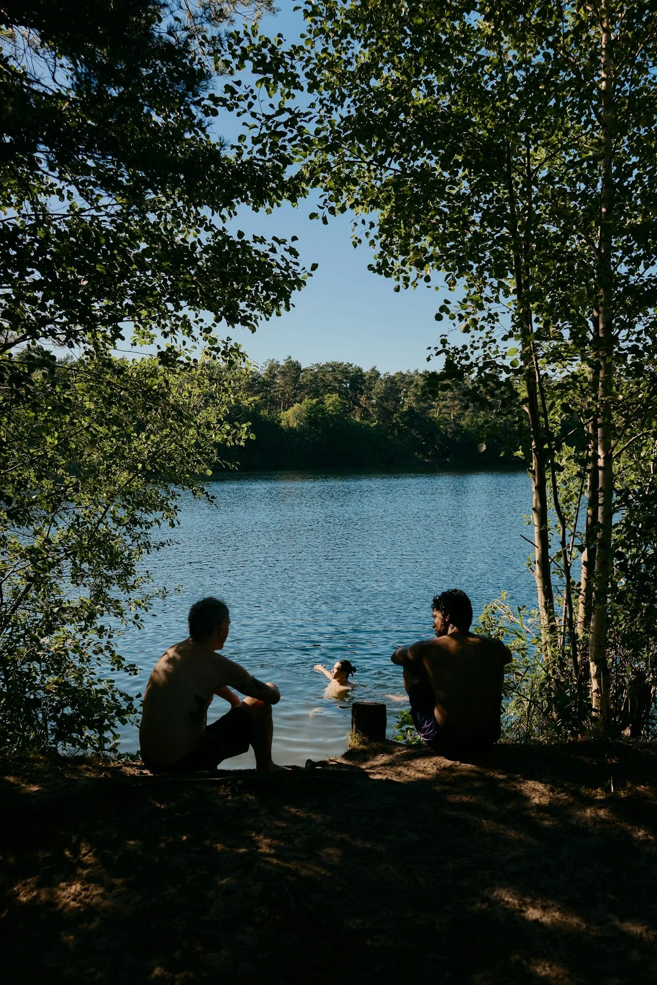 Two men sitting by the edge of a lake, looking at a child swimming in the water, with trees and clear blue sky in the background.