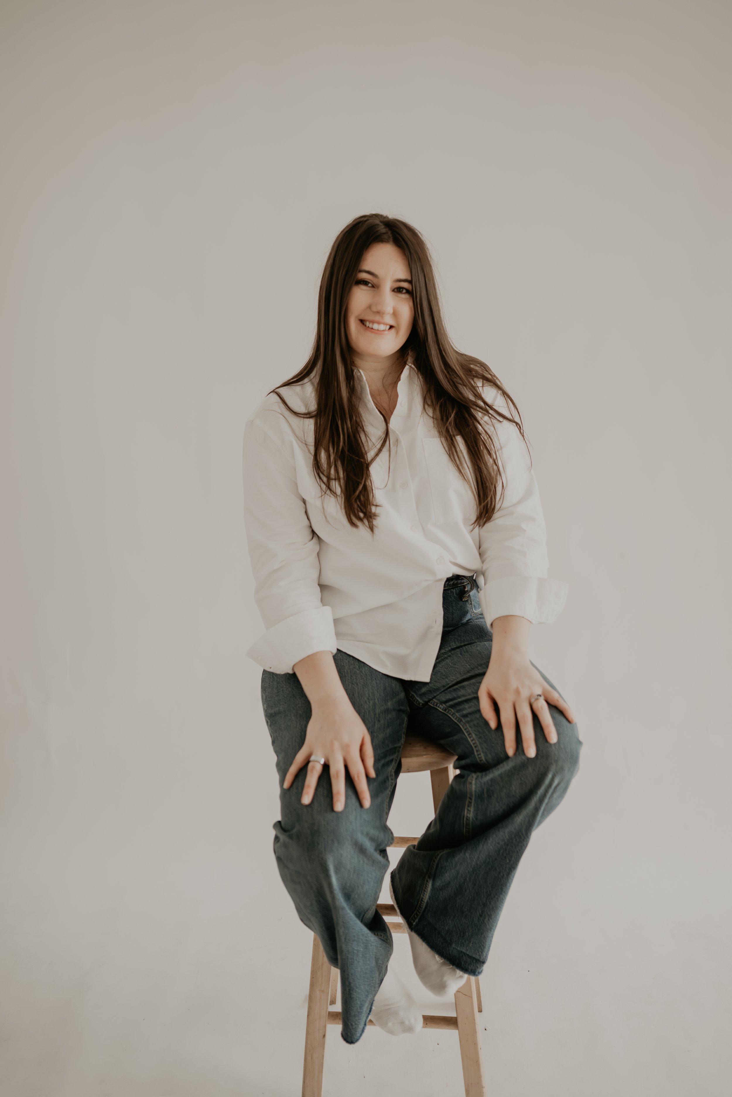 A woman with long brown hair, wearing a white shirt and dark jeans, sitting on a wooden stool against a plain off-white background, smiling lightly at the camera.