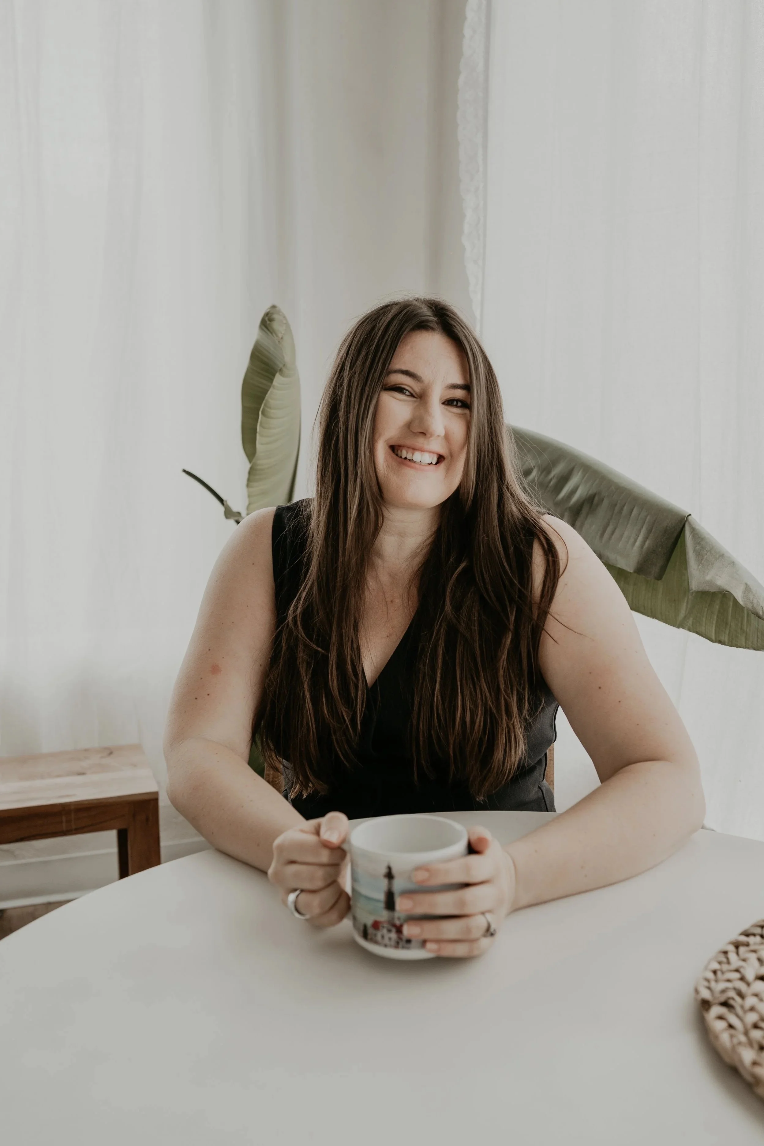 A woman with brown hair smiling while holding a mug, sitting at a white table in a bright room with white curtains and leafy plants in the background.