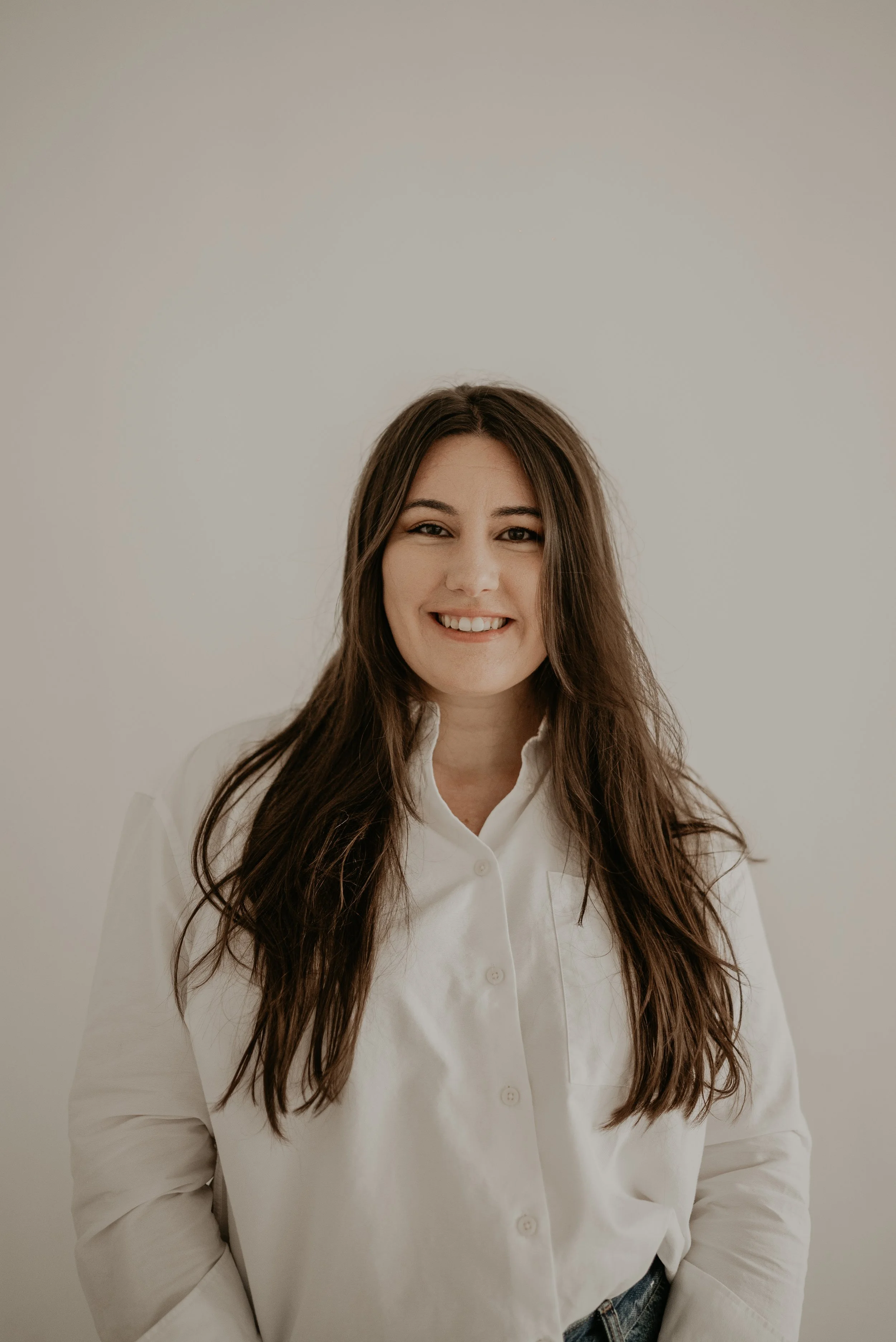 A woman with long brown hair, smiling, wearing a white button-up shirt, standing against a plain, light-colored background.
