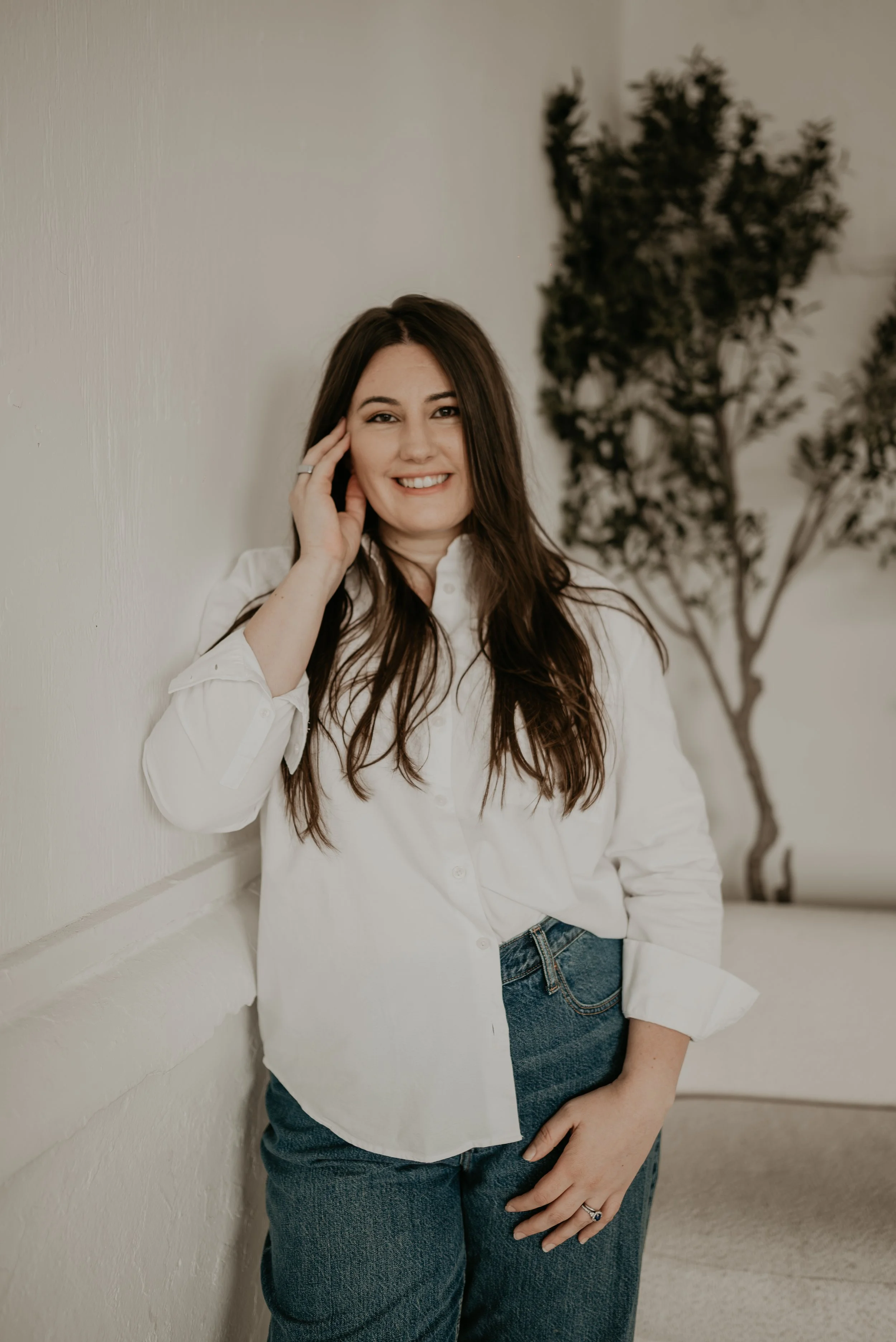 A woman with long brown hair and a bright smile, dressed in a white shirt and blue jeans, leaning against a white wall with a potted plant in the background.