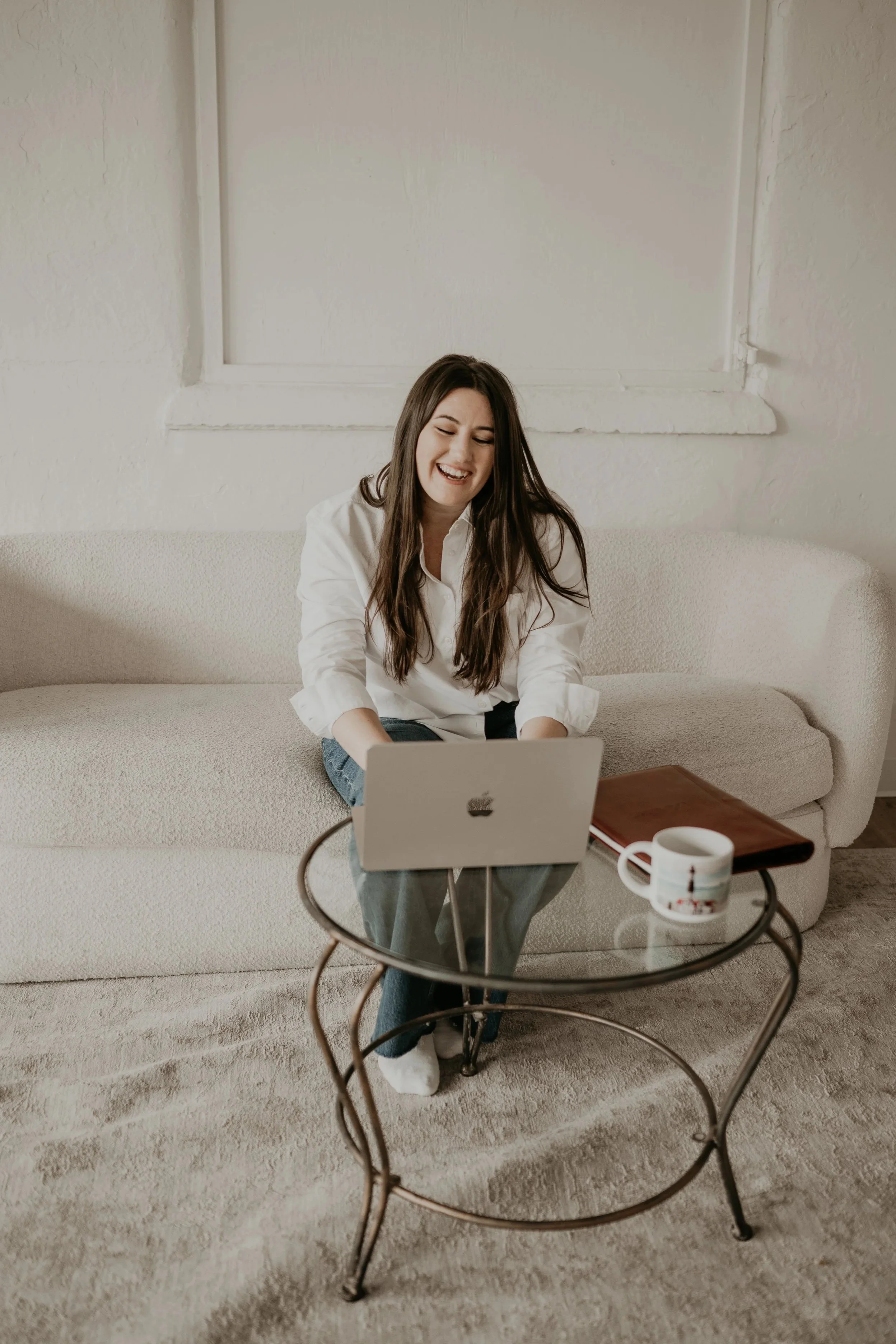 A woman sitting on a beige sofa, laughing, while working on her laptop placed on her lap. She is in a living room with a small round glass table in front of her, holding a white mug, a book, and a computer mouse. The room has a window with no curtains behind her.