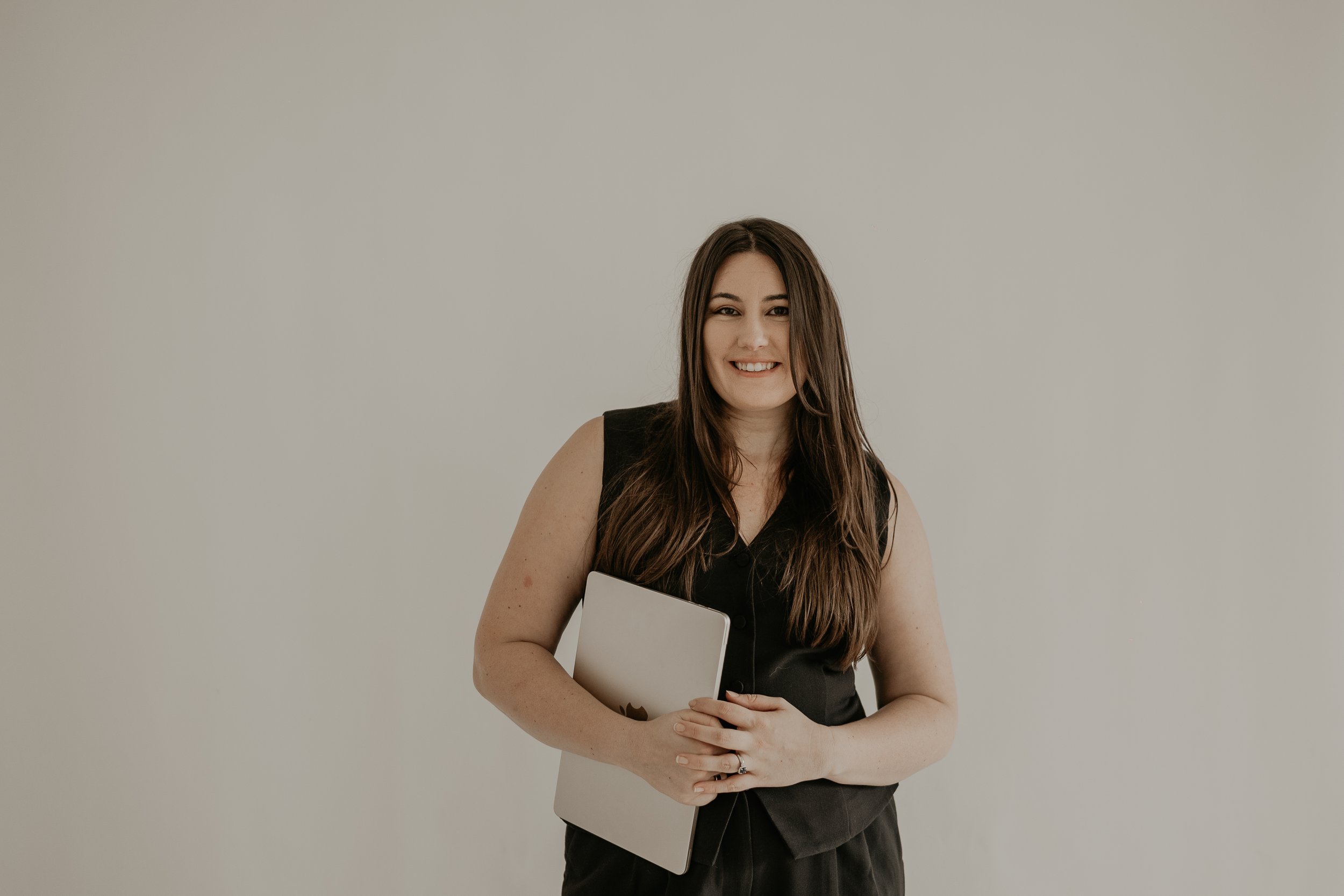 A young woman with long brown hair, smiling, holding a silver laptop and standing against a plain light gray background.
