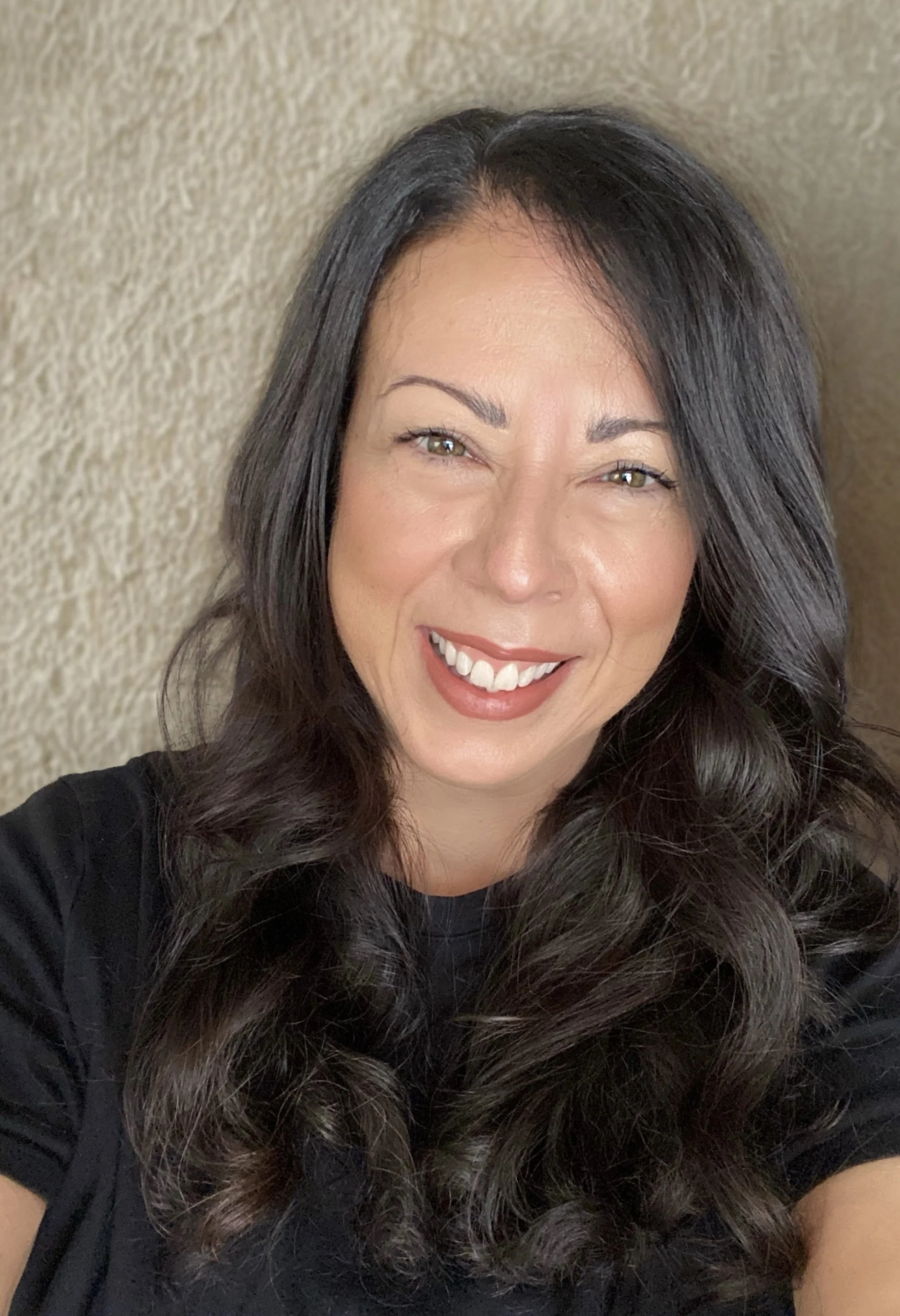 A woman with long, dark, curly hair and light skin smiling against a tan textured wall.