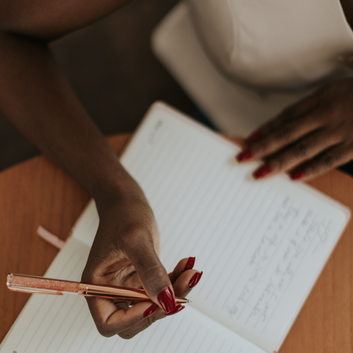 A person with dark skin and red painted nails writing in a lined notebook with a rose gold pen, sitting at a wooden table.
