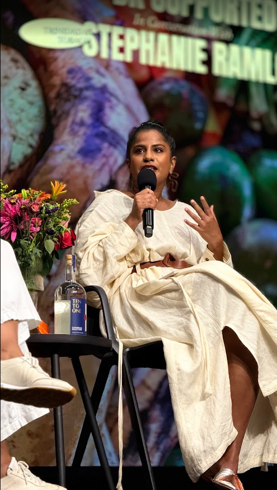 A woman sitting on a chair, speaking into a microphone during an event with a blurred colorful background and a large sign with her name, Stephanie Ram. There are flowers and a bottle of hand sanitizer on a small table next to her.