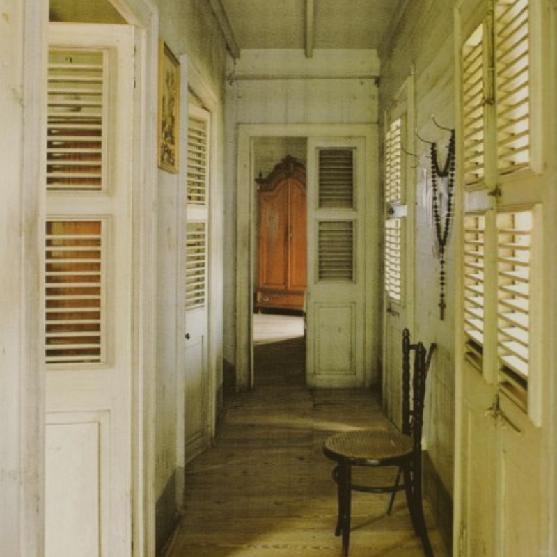 Interior hallway of an old house with shuttered windows, wooden floors, a wooden wardrobe visible through an open doorway, a wooden chair, and rosary beads hanging on the wall.