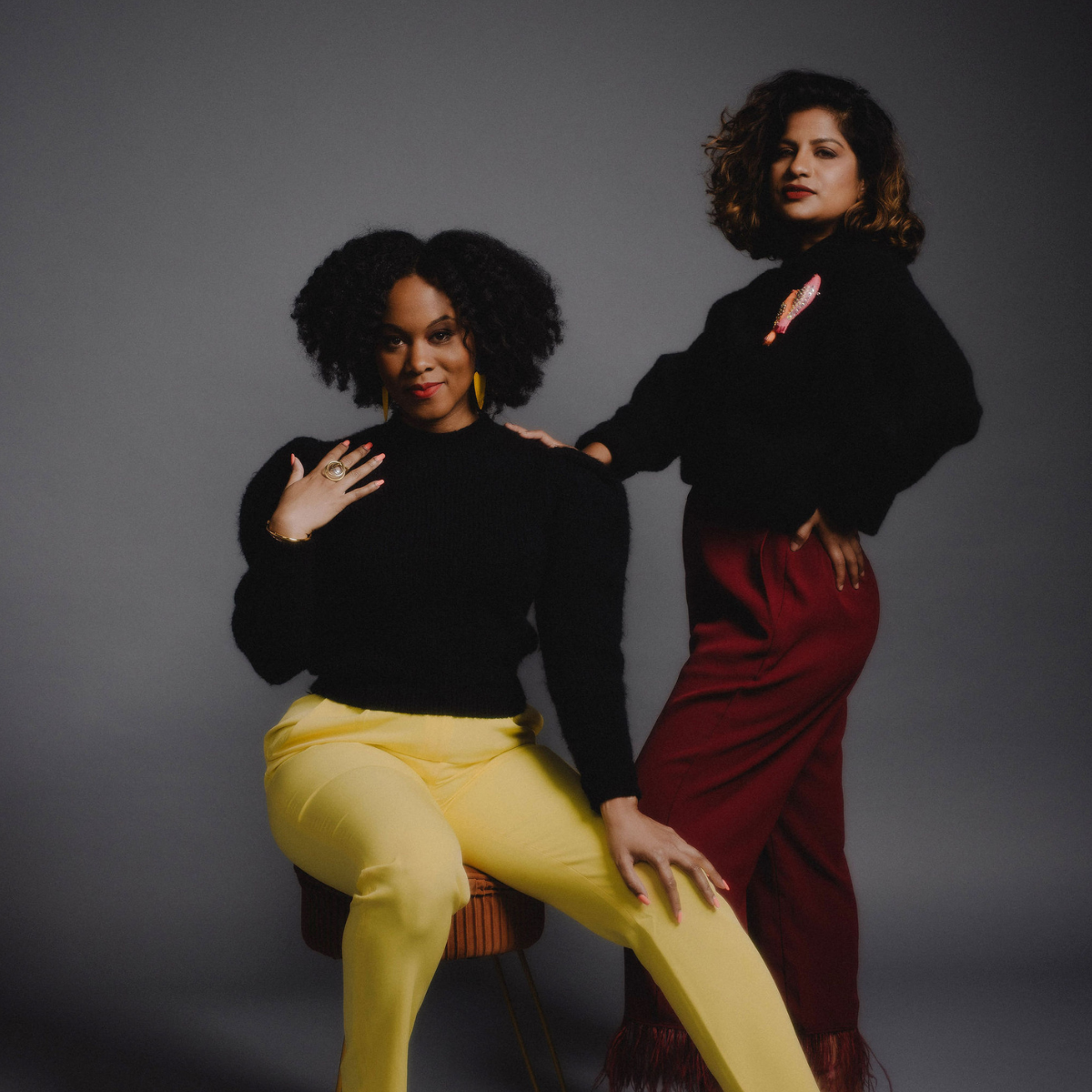 Two women with curly hair posing against a plain gray background. The woman on the left is seated, wearing a black sweater and yellow pants, with yellow earrings and a ring. The woman on the right stands with hand on her hip, wearing a black sweater with decorative patches and red high-waisted pants.