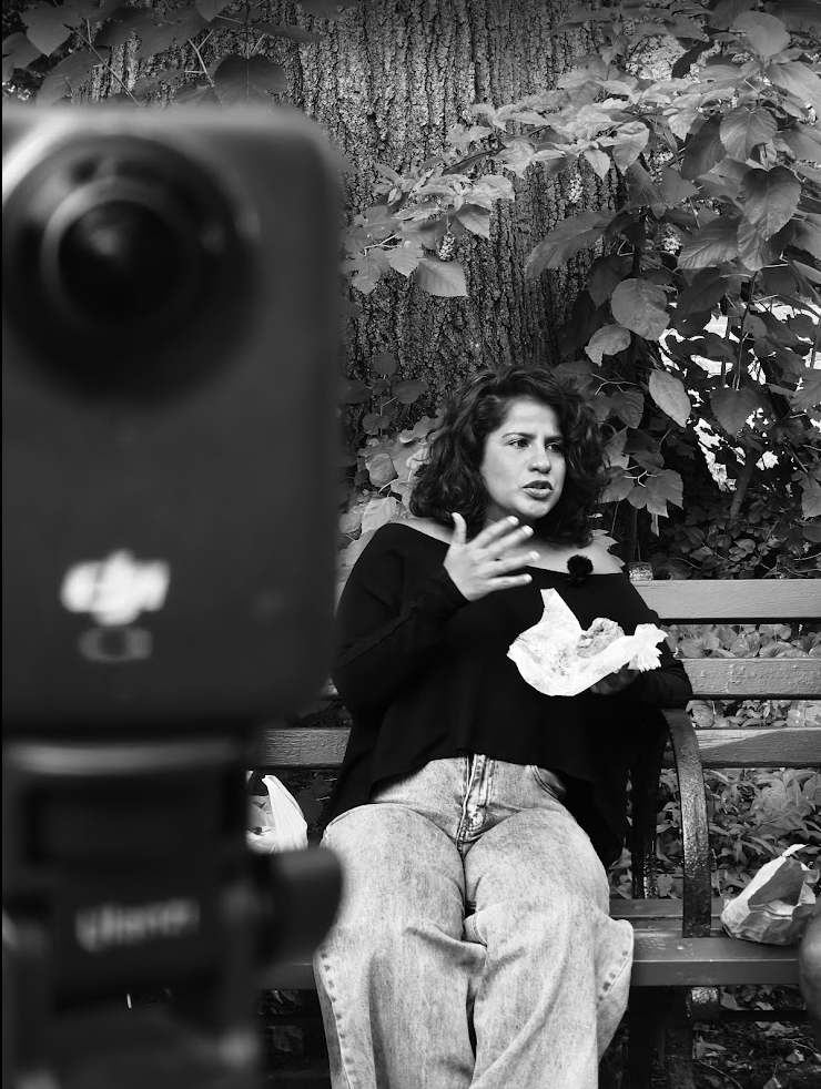 A woman sitting on a park bench, speaking and holding a piece of paper, with a large tree and leafy plants in the background, as a camera captures her in black and white.