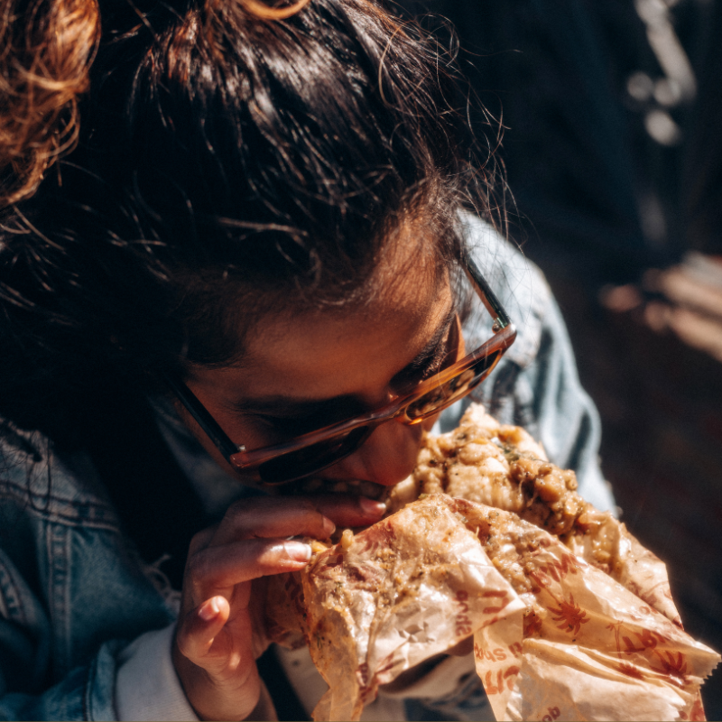Woman in sunglasses eating a large a doubles from Queens NY wrapped in paper.