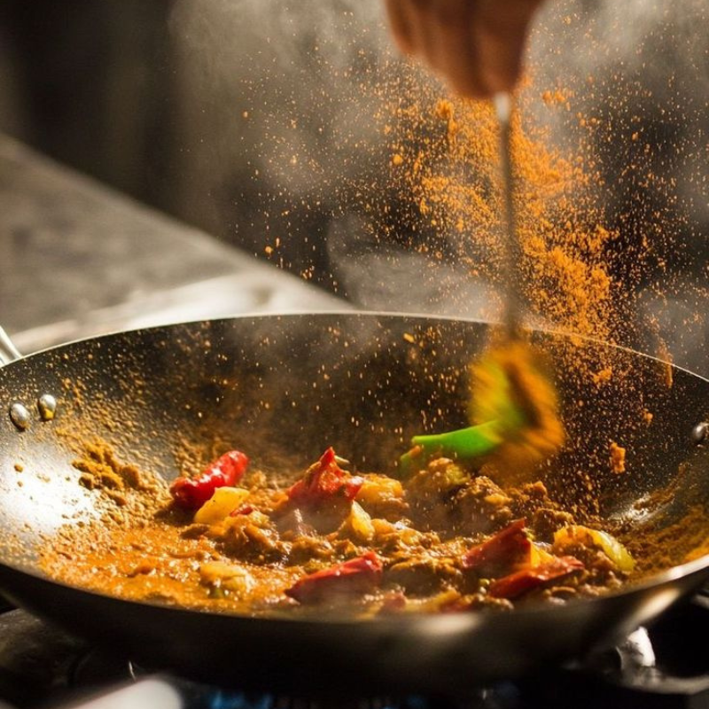 A person cooking spicy stir-fry with red and yellow peppers in a wok, adding seasoning or spices.