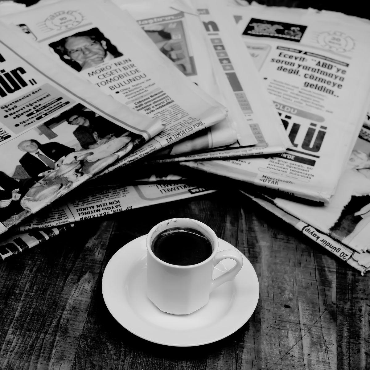 A white cup of black coffee on a saucer placed on a wooden table, with a pile of newspapers scattered behind it.
