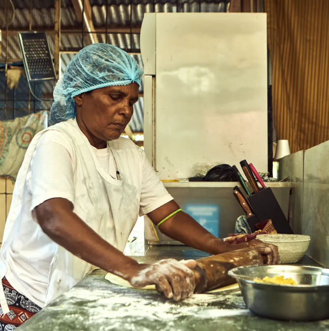 Woman wearing a hairnet and white apron rolling roti on a floured surface in a rustic kitchen in Trinidad. SHot by Marlon James