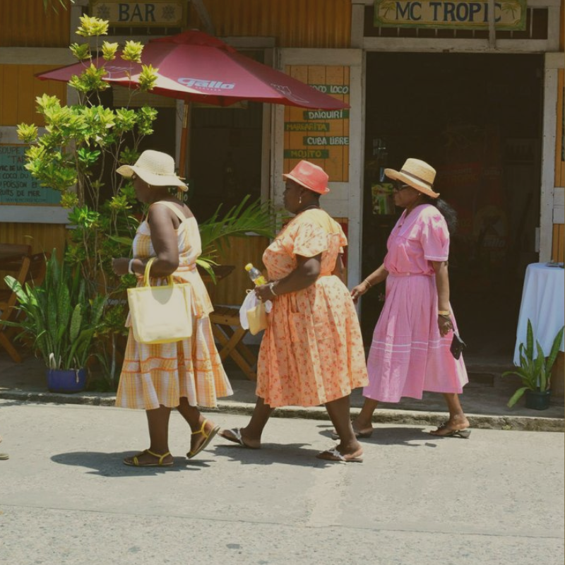 Three women walking on a street near a restaurant or bar, wearing colorful dresses and wide-brimmed hats, carrying handbags, with potted plants and a large red umbrella in the background.