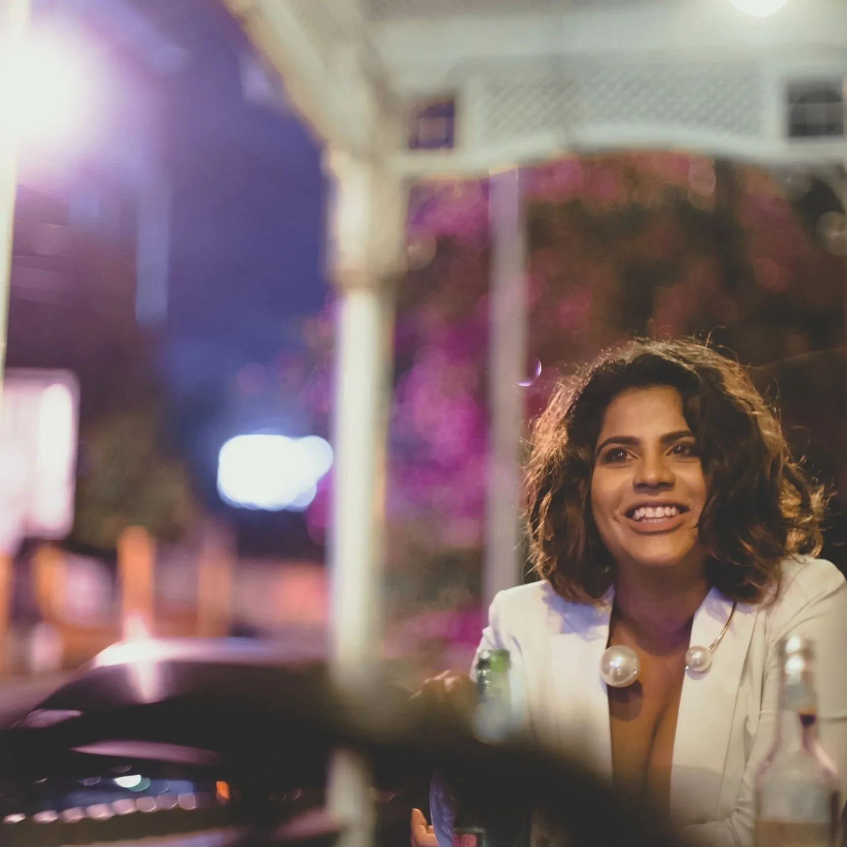 A woman with curly hair and a white blazer smiling in a restaurant or bar on a night out with colorful lights in the background.