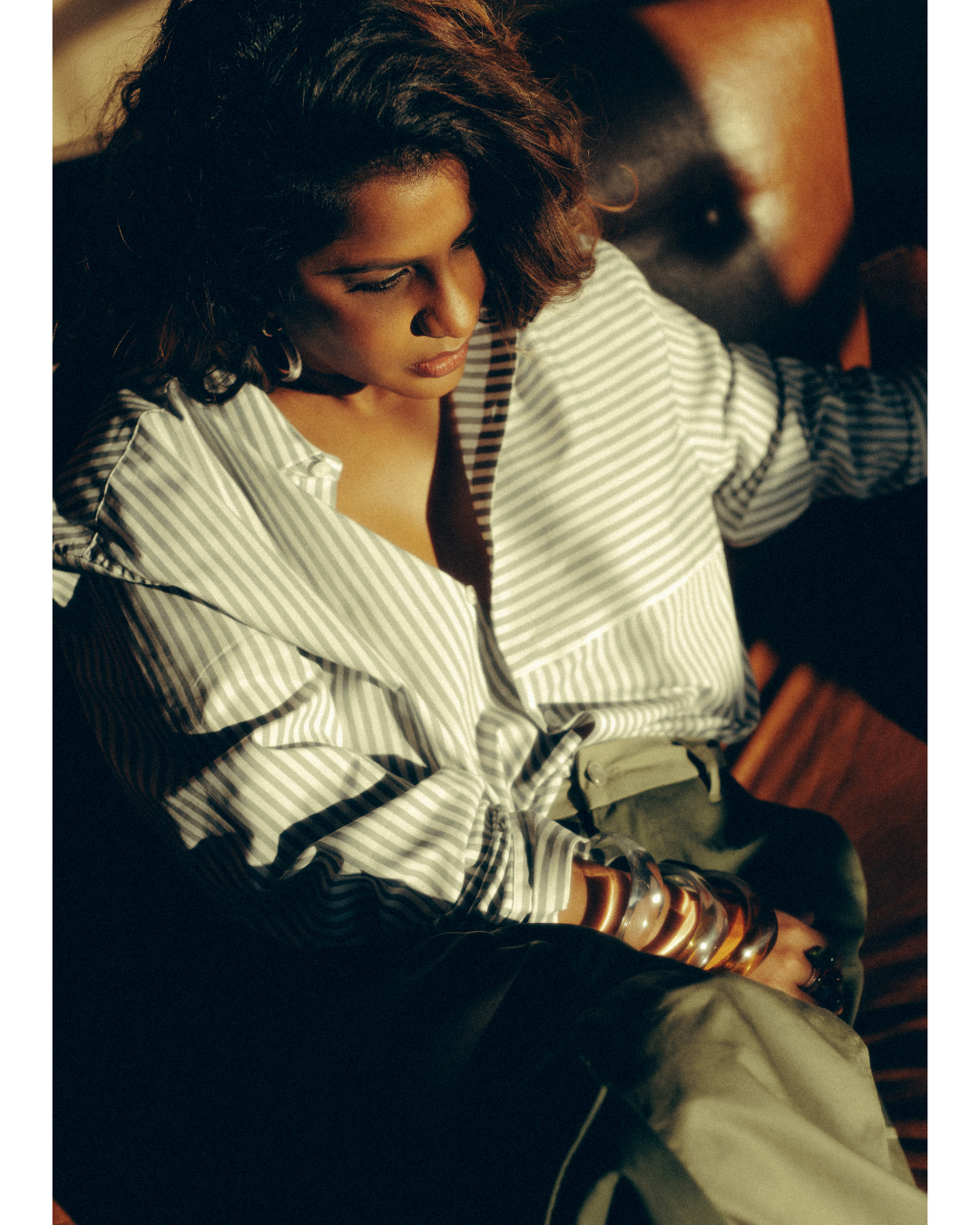 A woman with curly brown hair wearing a striped shirt seated indoors with sunlight on her face, looking downward.