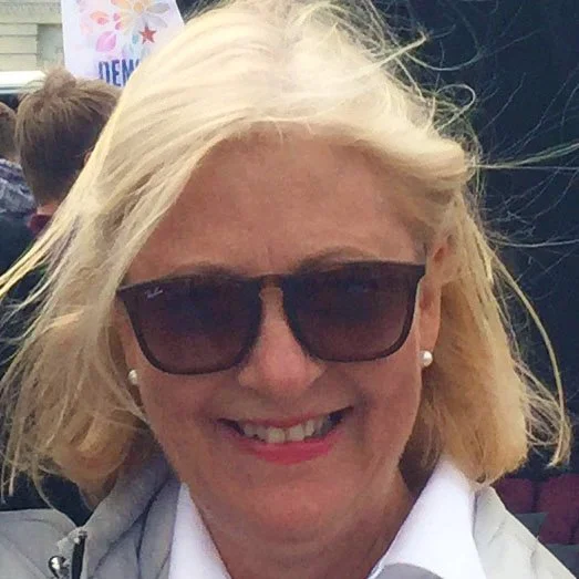 Smiling woman with blonde hair, wearing sunglasses, pearl earrings, and a white shirt.
