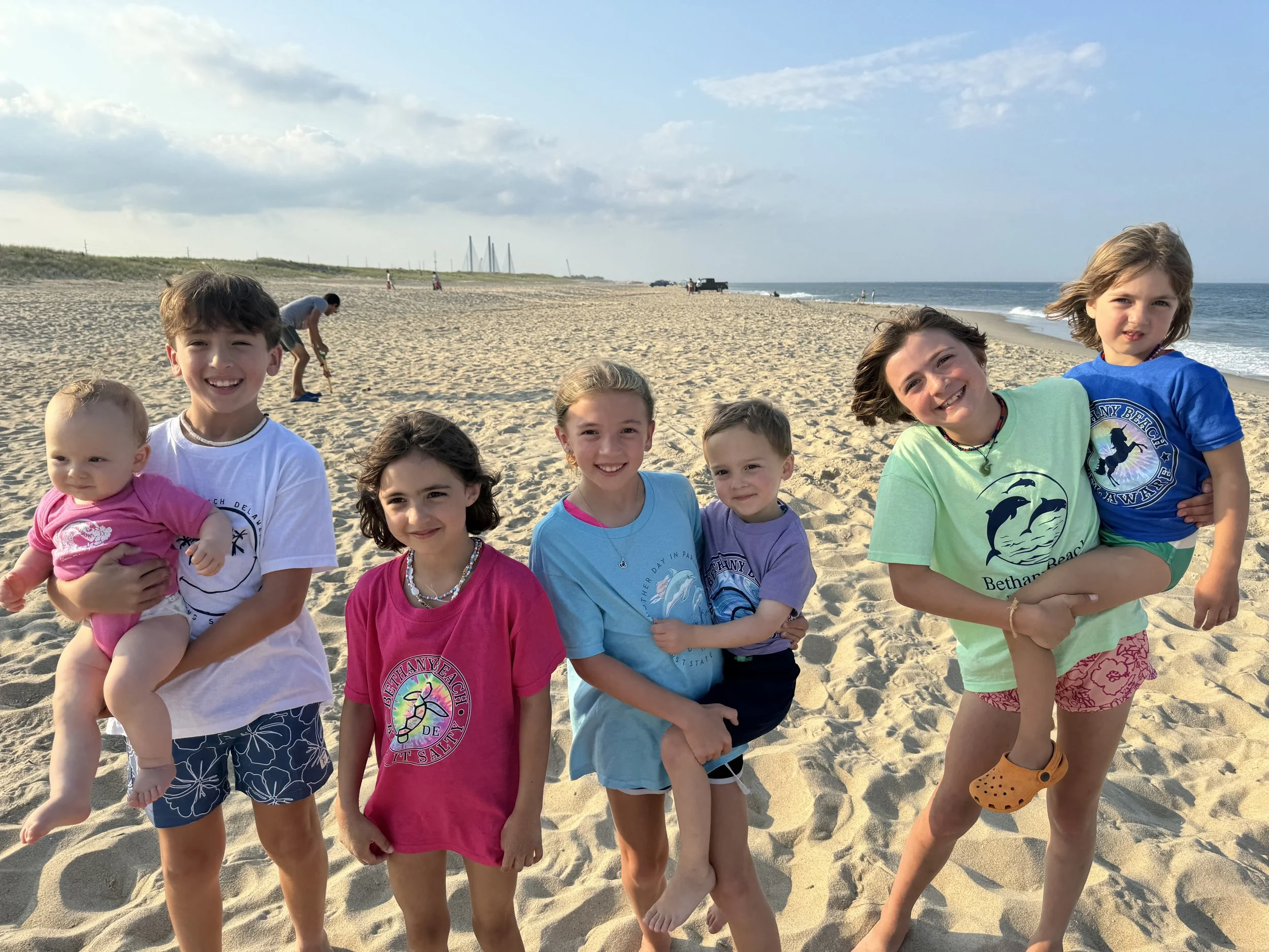 Group of children and an adult woman smiling and posing for a photo on a sandy beach with the ocean and cloudy sky in the background