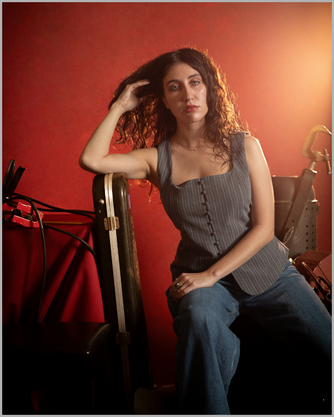 A woman with curly brown hair sitting against a red background, looking at the camera with a neutral expression, wearing a sleeveless gray pinstripe top and blue jeans, with electrical equipment and tools around her.