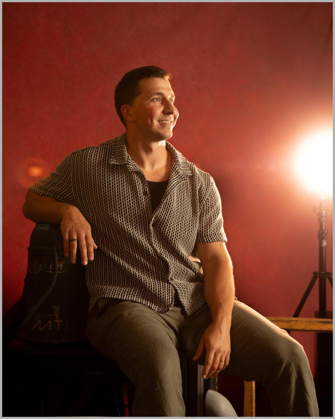 A smiling man sitting sideways on a chair with his left arm resting on a skateboard. The background is a red wall, and there is a bright light on the right side of the image.