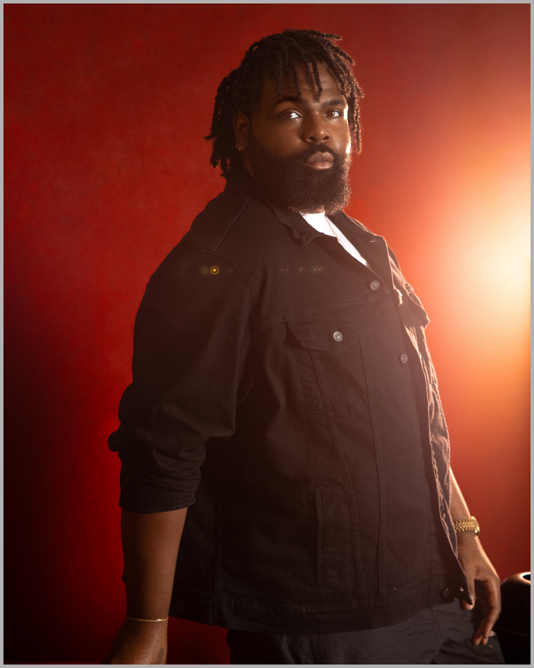 A man with dreadlocks and a beard wearing a black denim jacket stands in front of a red background with lighting creating a glowing effect on his right side.