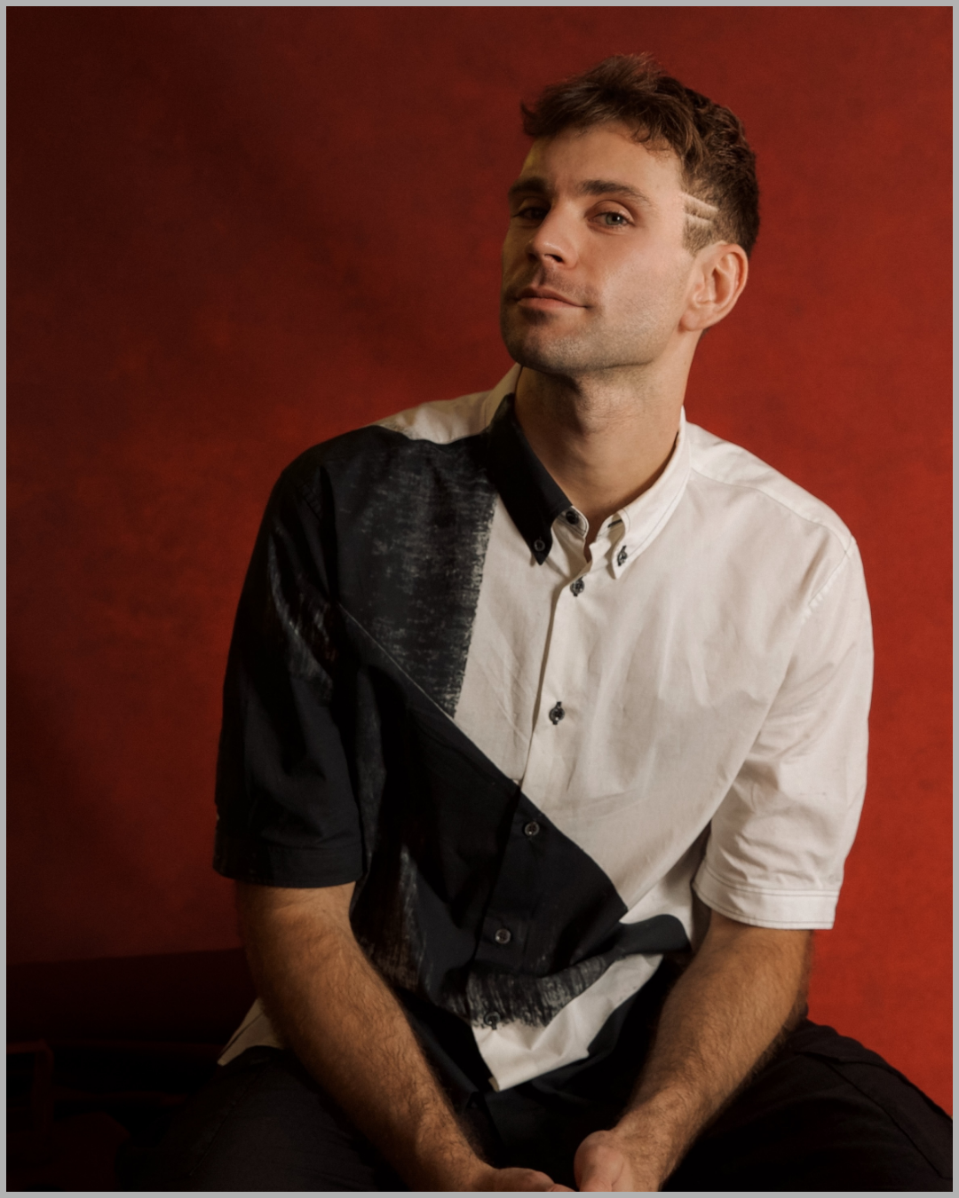 A young man with short, light brown hair and a slight beard, sitting against a red textured background. He is wearing a short-sleeved, button-up shirt with black and white patterns and has a confident expression.