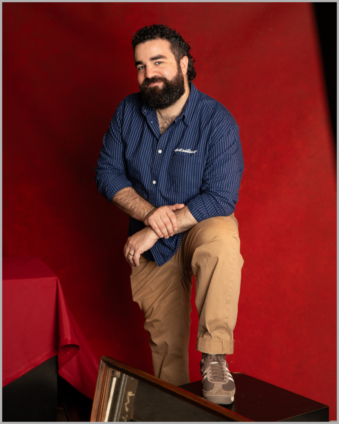 A man with a beard and curly hair, wearing a blue striped shirt and beige pants, is posing against a red background. He is leaning on one knee with his hands clasped, one foot on a black step, and smiling.